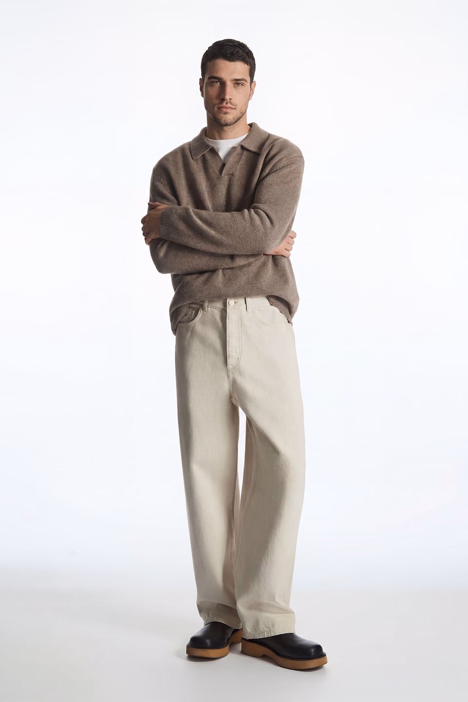 Nick, a Botika Ai fashion model is standing in a white studio background wearing a light brown polo sweater and oversized khakis.