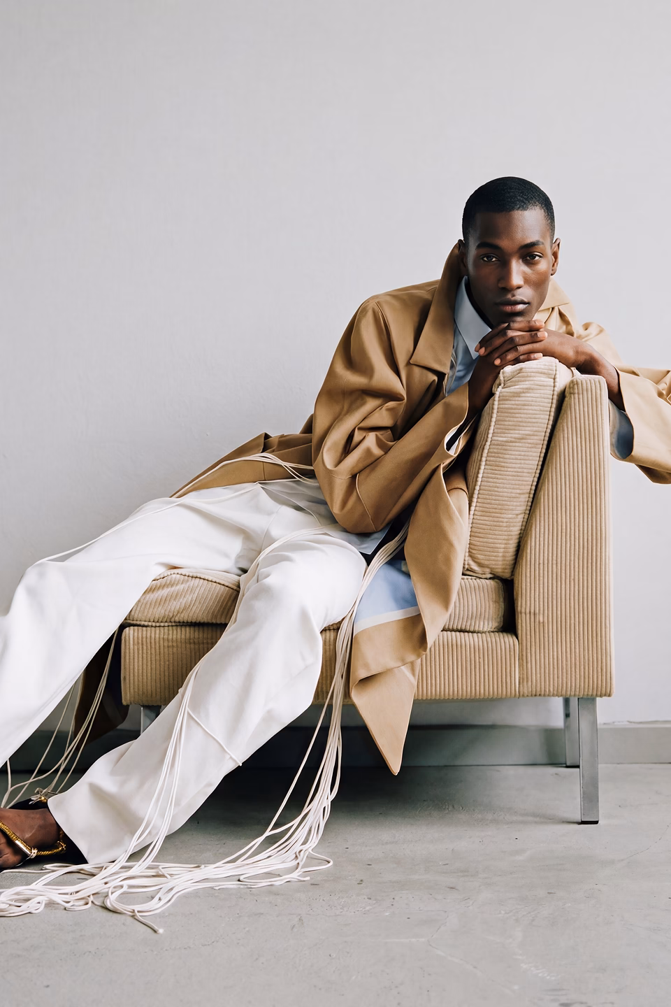 Black male AI fashion model named Tre is lounging on a chaise wearing white pants a powder blue collard shirt and camel overcoat.