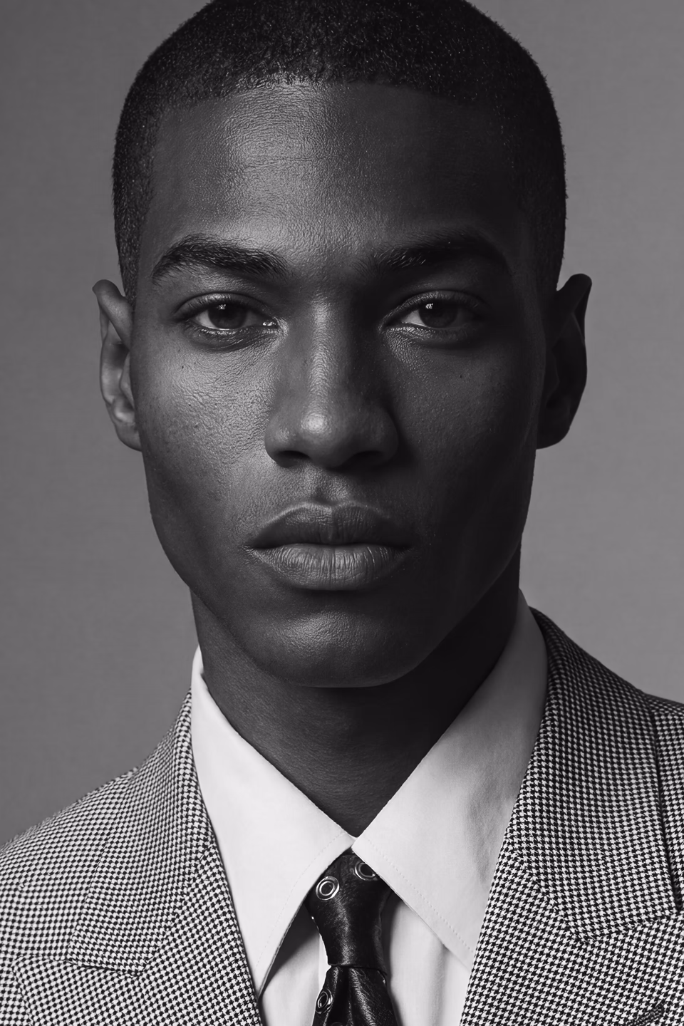 A black and white headshot of Tre, a black male AI fashion model wearing a houndstooth blazer, white collared shirt and dark patterned tie.