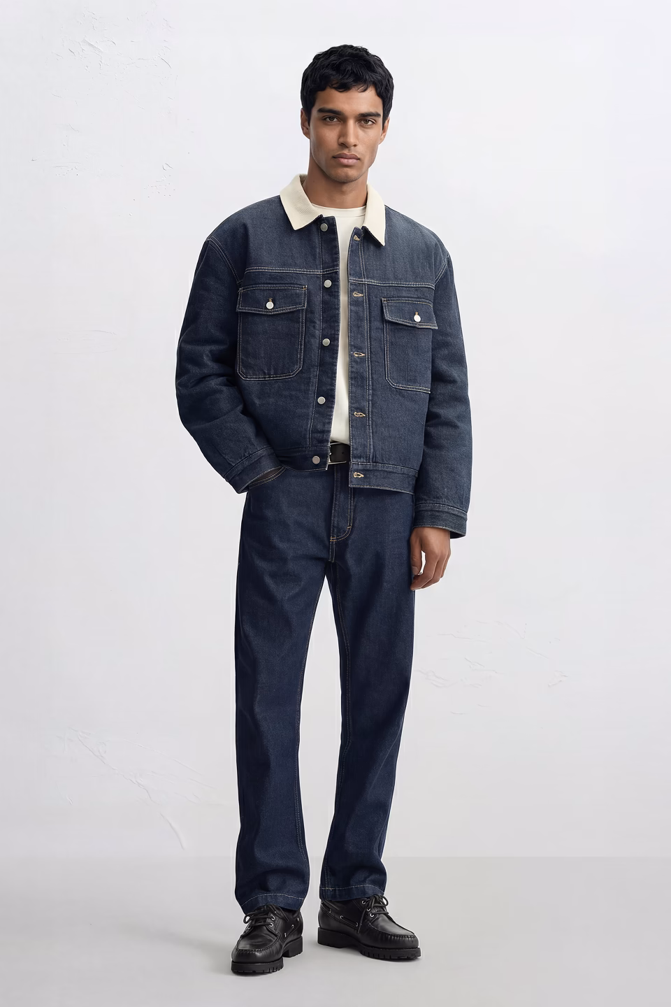 Kai, a Botika male AI fashion model standing wearing a jean jacket with a white collar, and jeans with a white studio background.