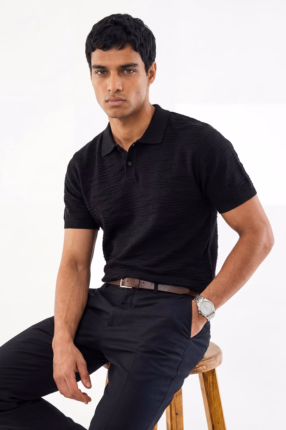 Botika male AI fashion model Kai sitting on a stool wearing a dark polo and matching dress pants.