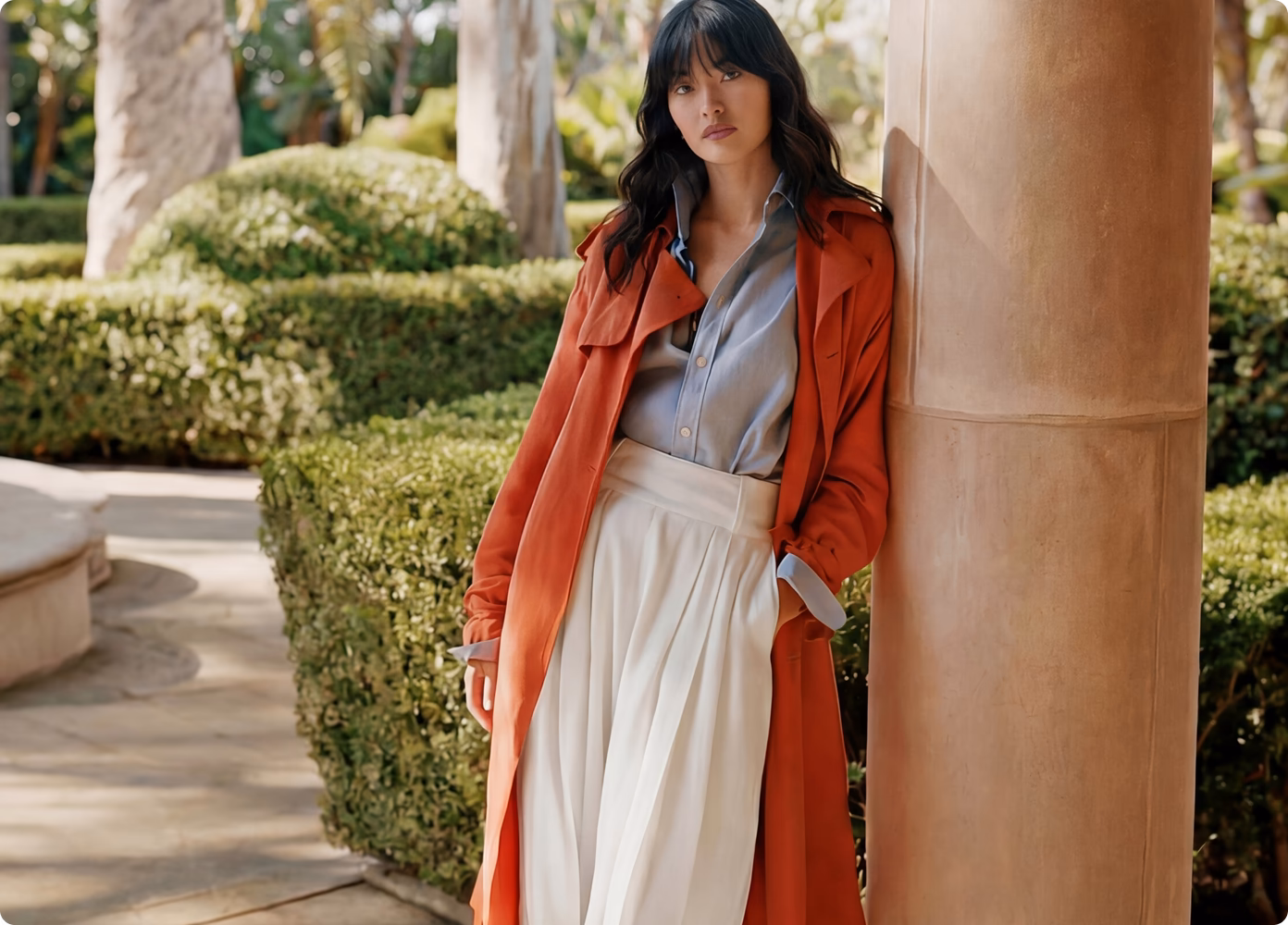 A female Asian AI fashion model leaning against a column in a garden wearing white pants, a light blue button down shirt and burnt orange trench coat.