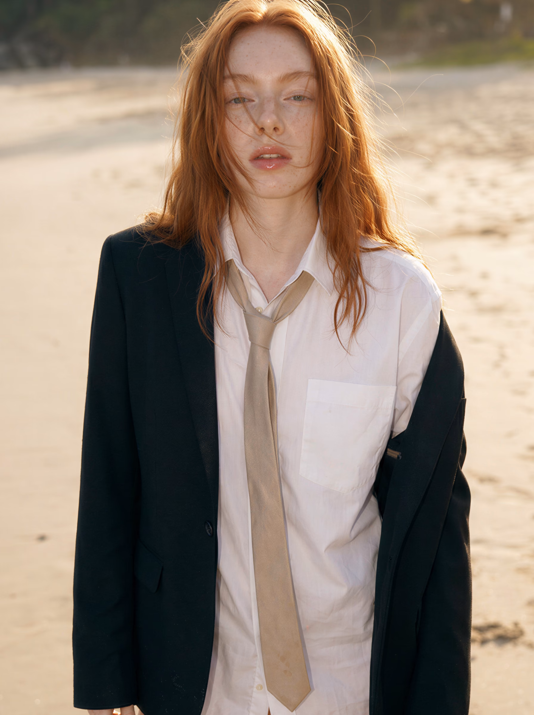 Woman with long red hair wearing oversized black blazer and white shirt with beige tie standing on sandy beach