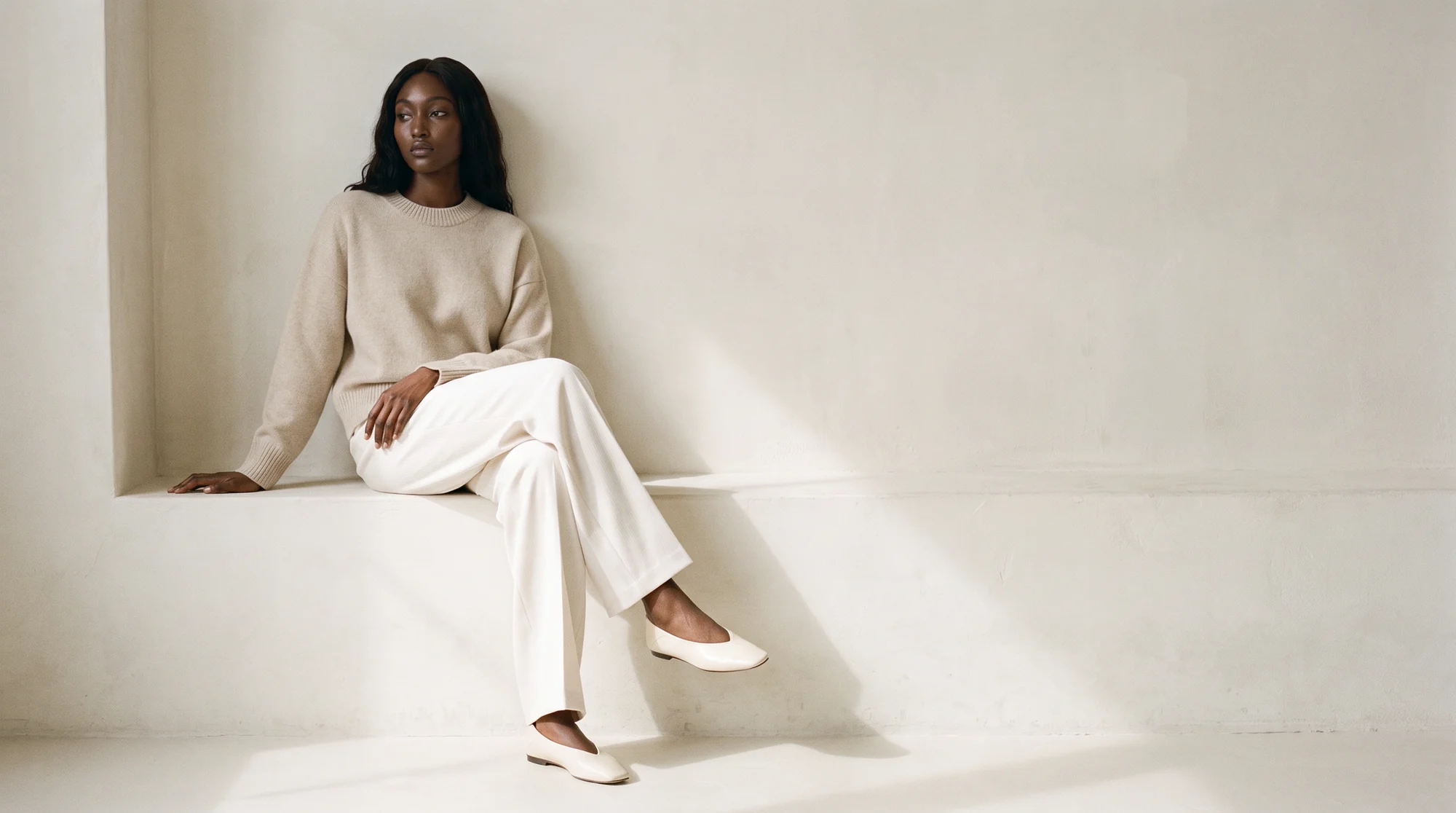 Woman seated against a cream wall in a knit sweater and ivory trousers, AI on-model editorial image