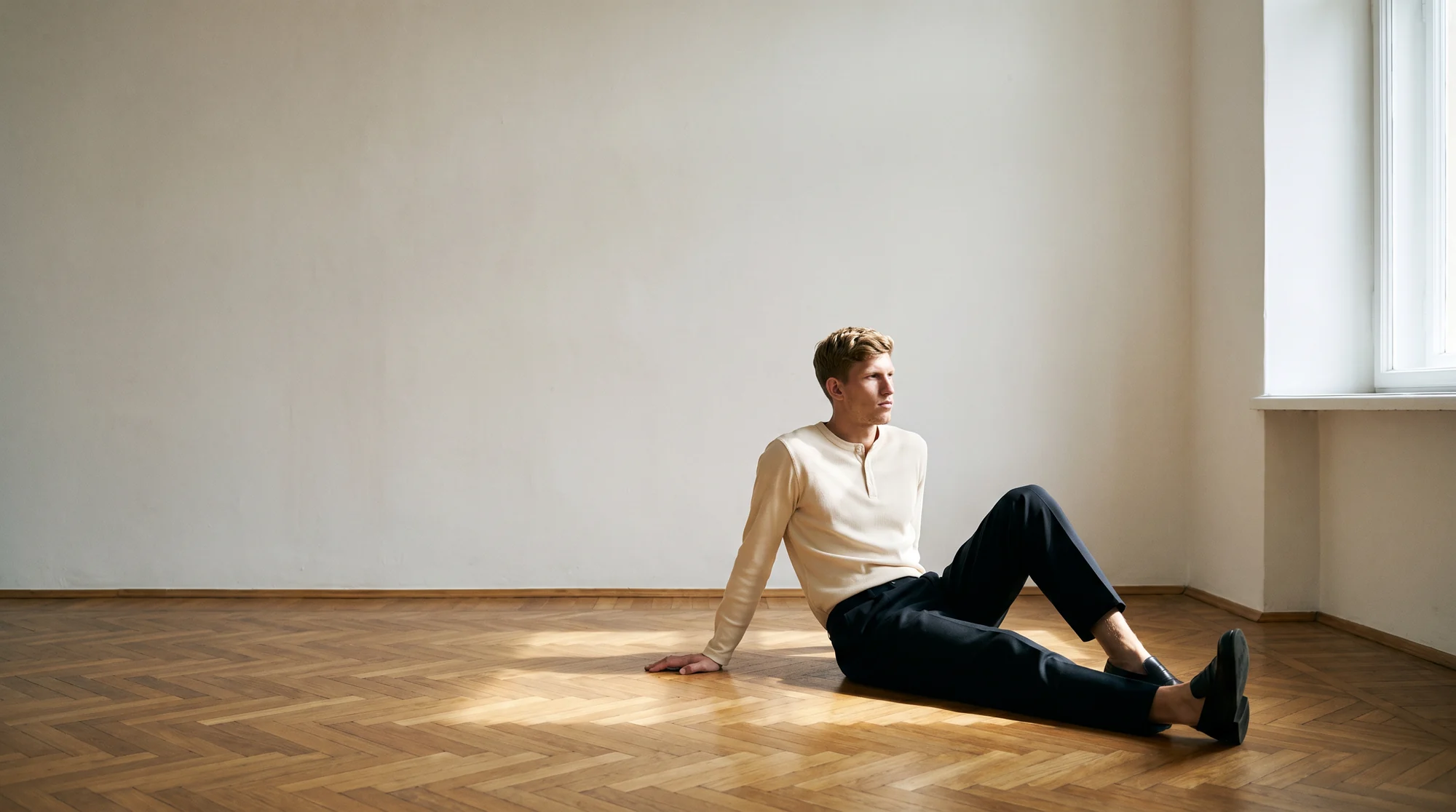Man seated on a concrete floor in a cream henley and black trousers, AI on-model editorial image
