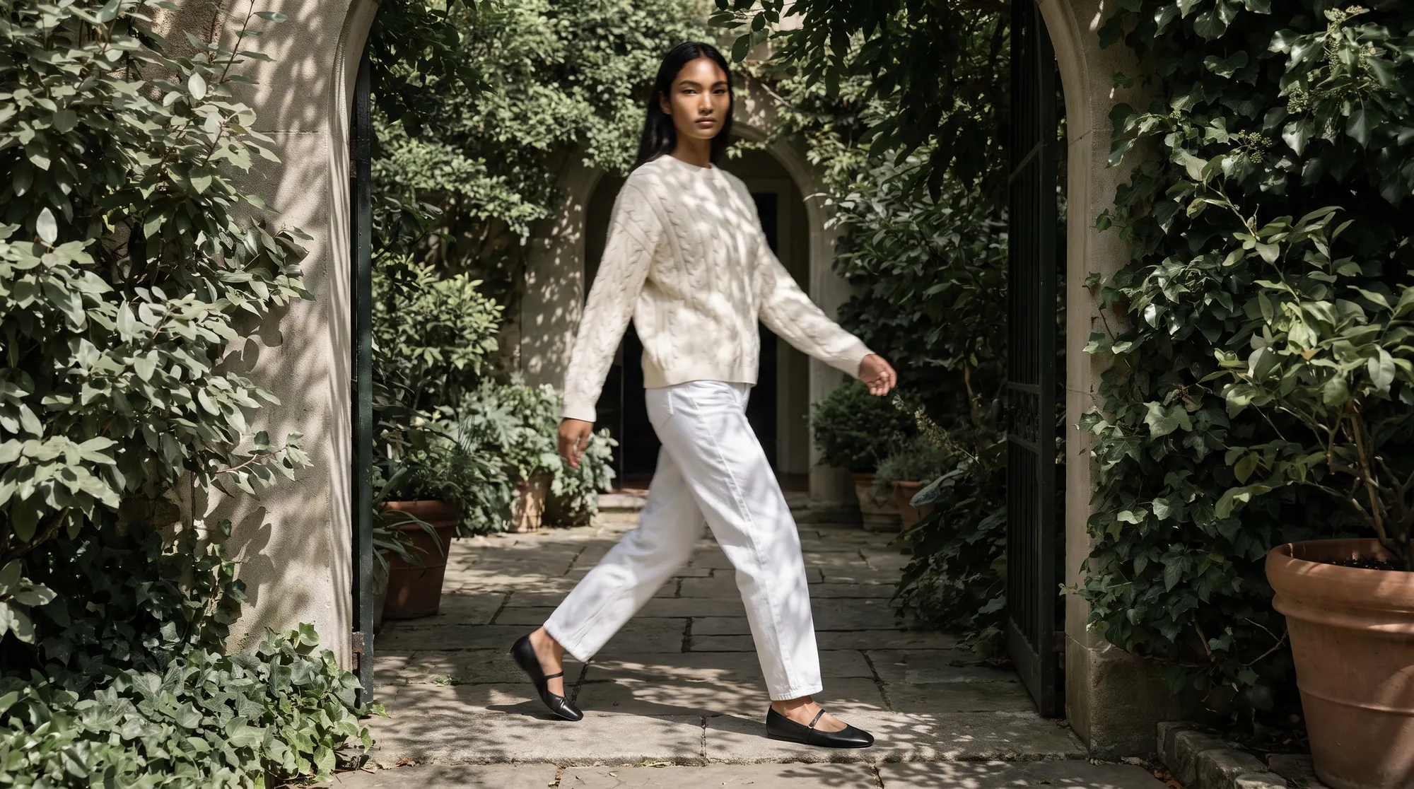 Model in a cream cable knit striding through a doorway into a courtyard dense with green foliage
