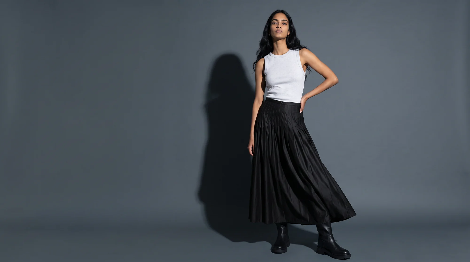 Model in a white top and black pleated skirt lit by hard flash against a slate charcoal sweep
