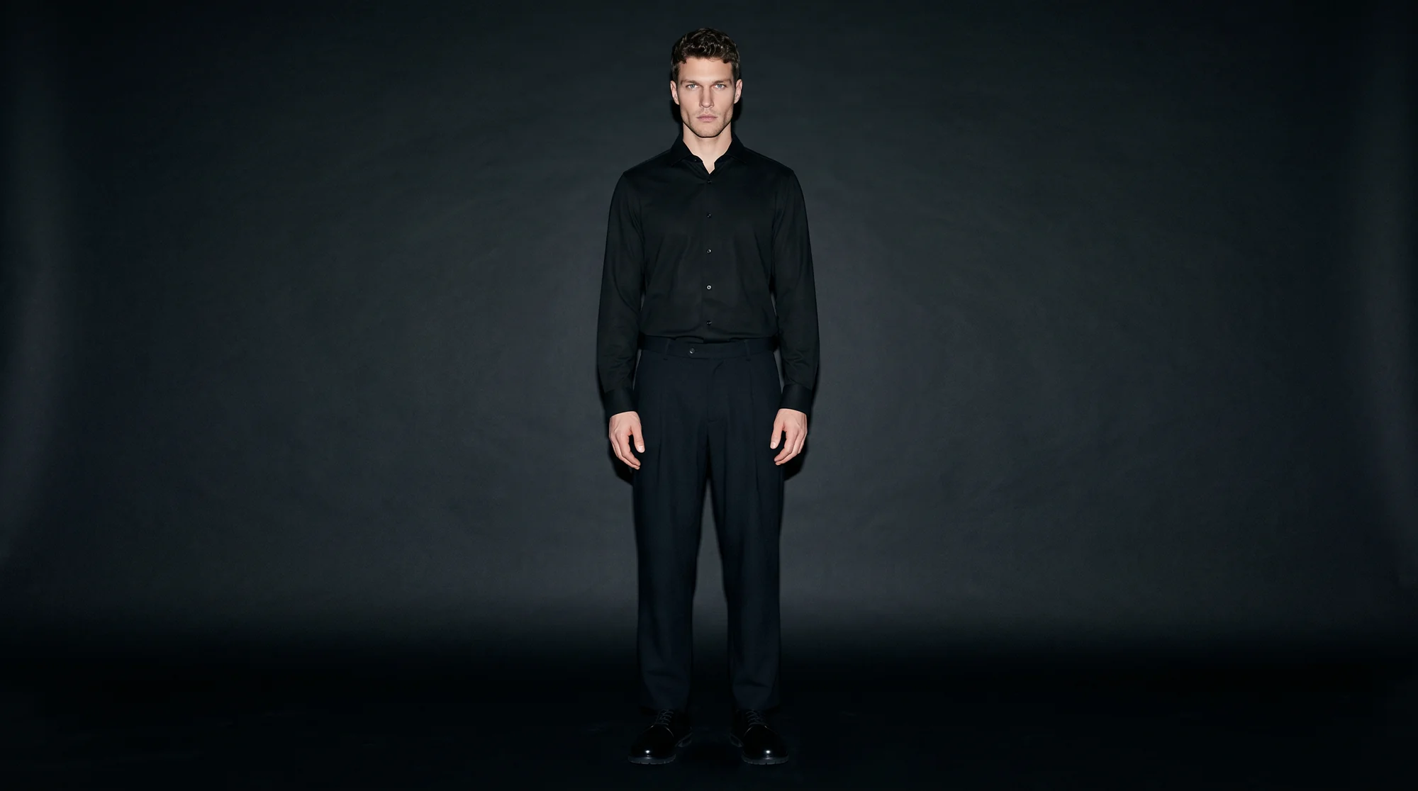 Man in a black shirt and trousers lit by hard flash against a deep charcoal studio backdrop