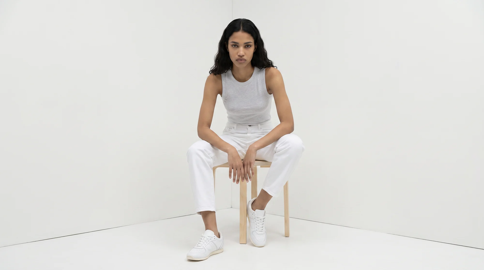 Model seated on a pale wooden stool in the corner of a white studio, dressed in white