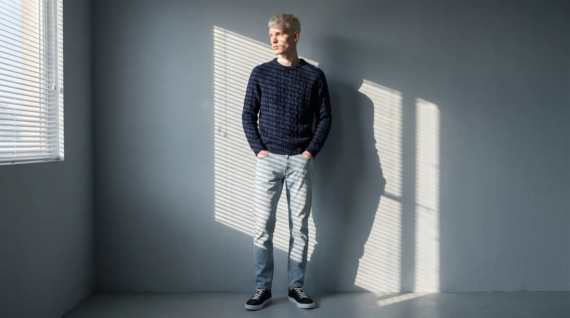 Man in a navy cable knit standing in a bare room as venetian blinds cast striped light across the wall