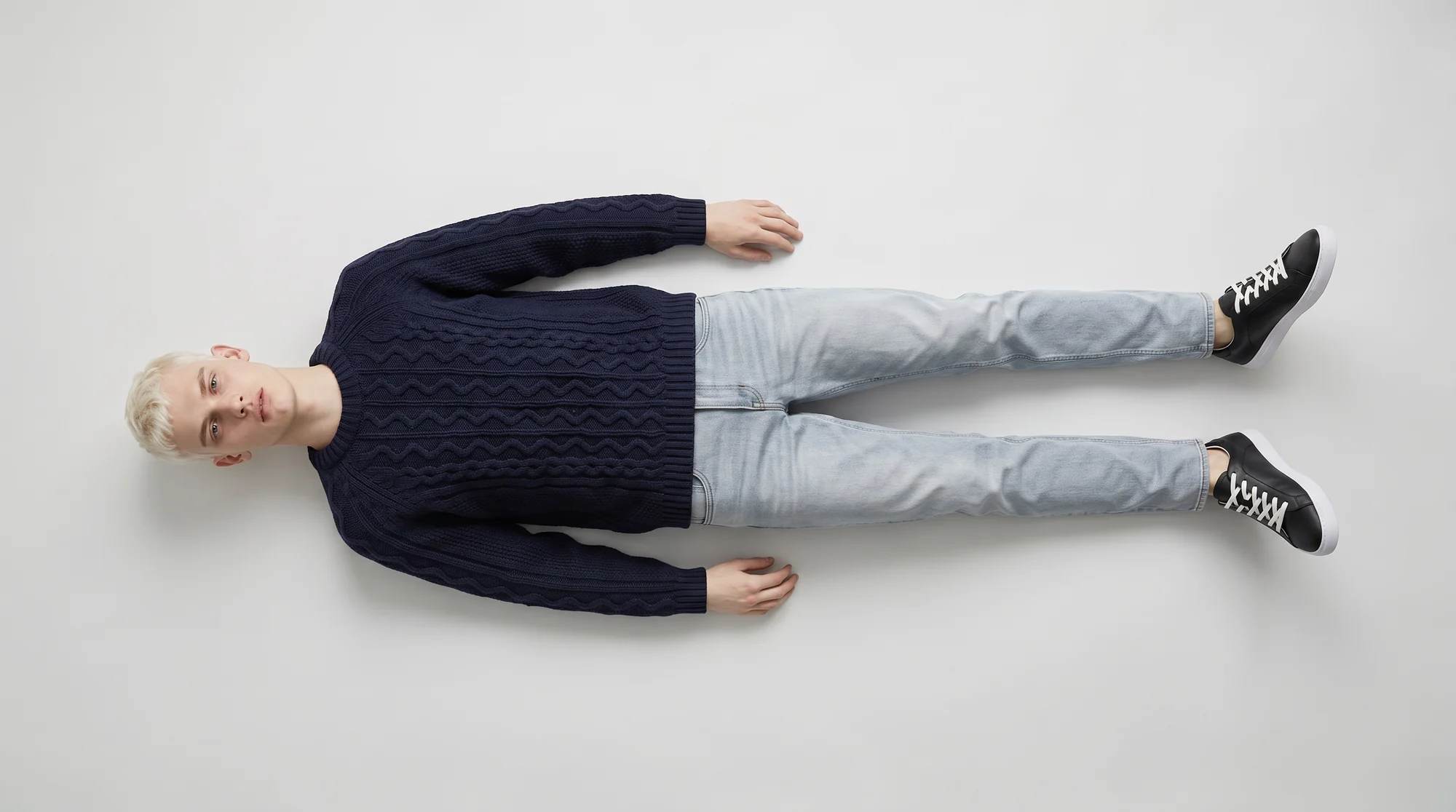 Overhead view of a man lying flat on a pale studio floor in a navy knit and light jeans