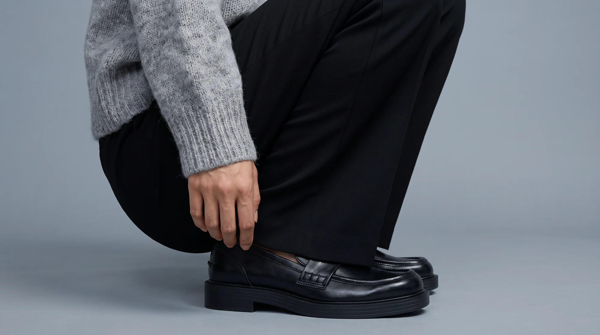 Tight crop on a grey knit sleeve cuff, hand and black trousers above a black leather loafer