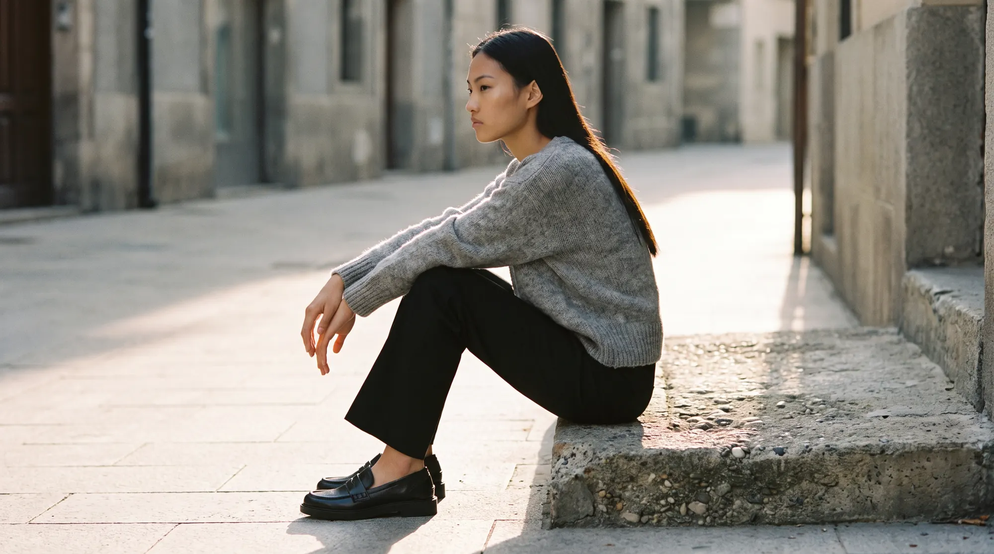 Model seated in profile on a low concrete step outdoors in warm late afternoon side light
