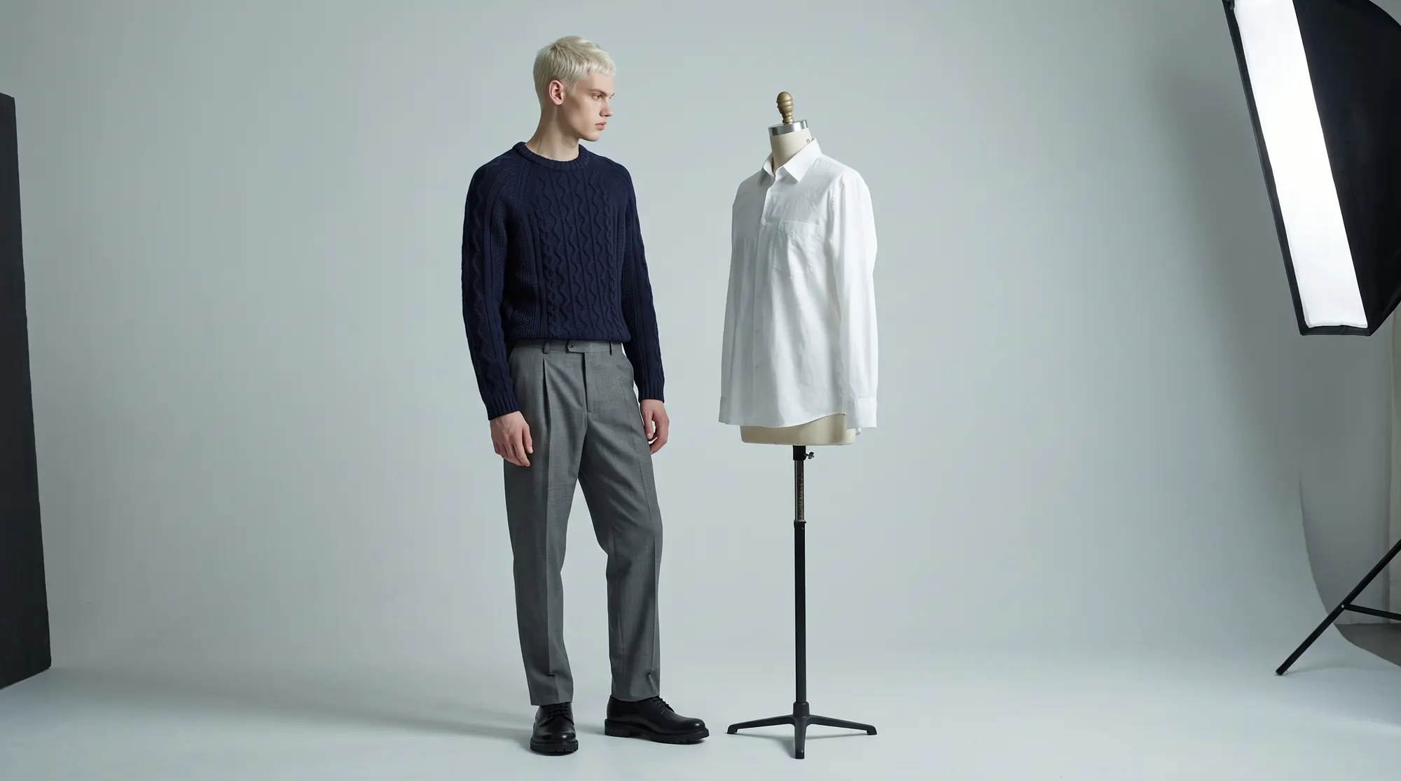 Man in a navy knit standing beside a tailor's dress form wearing a white shirt, softbox at the frame edge