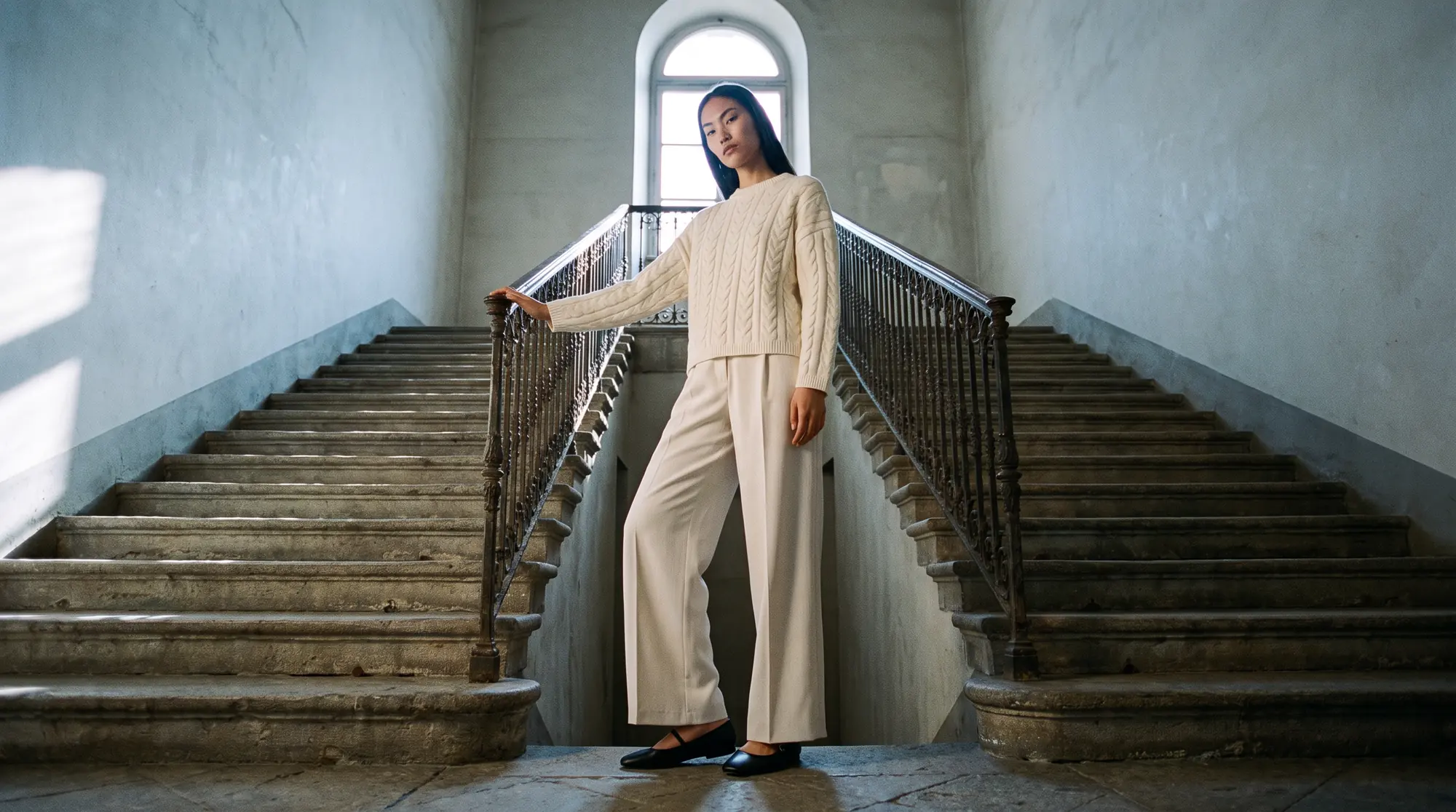 Model standing on the landing of a wide stone staircase where two flights divide, hand on the iron balustrade