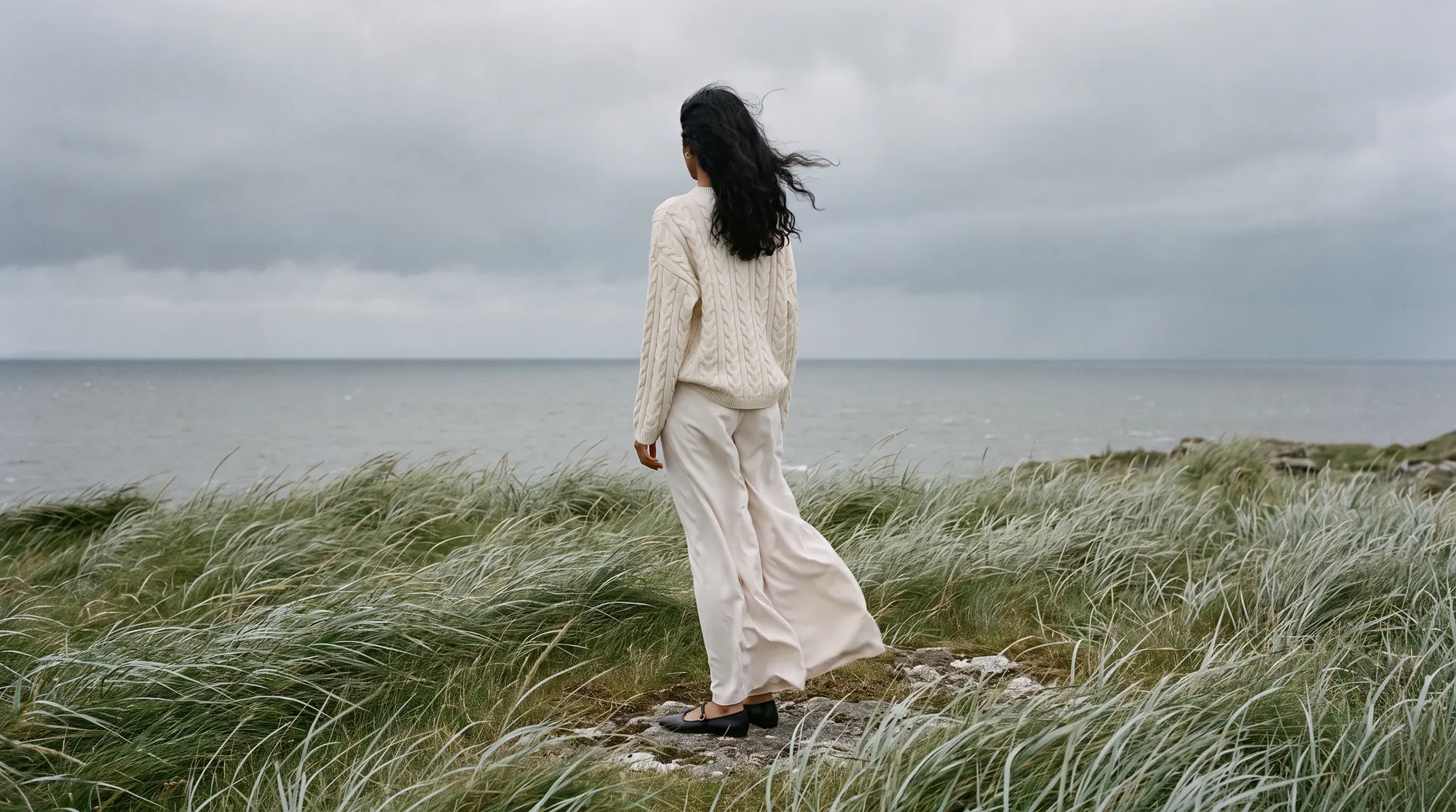 Model on a wild coastal headland with silver grass bending in the wind, looking out to a flat grey sea