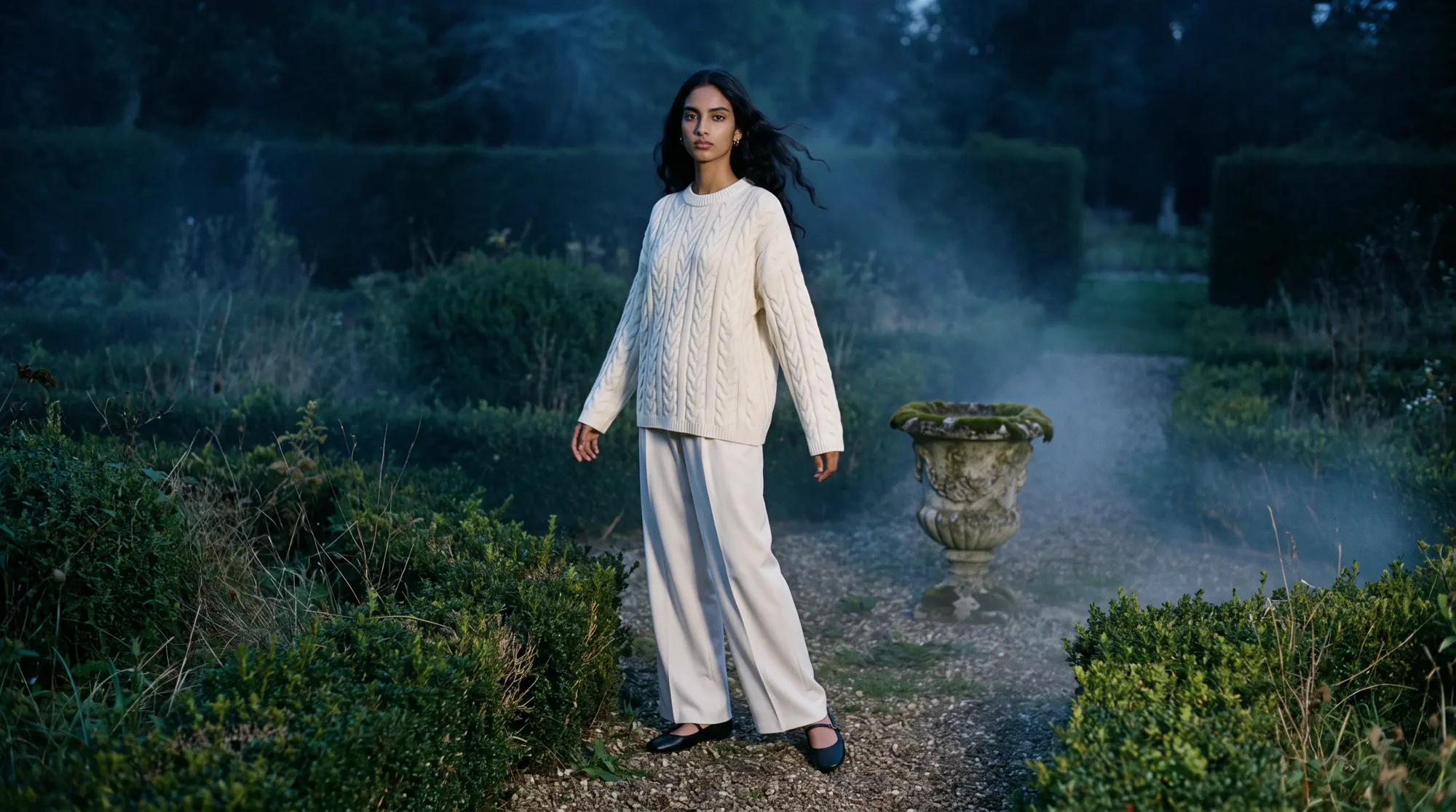 Model in a cream cable knit among the hedges of an overgrown formal garden at dusk, mist low over the gravel