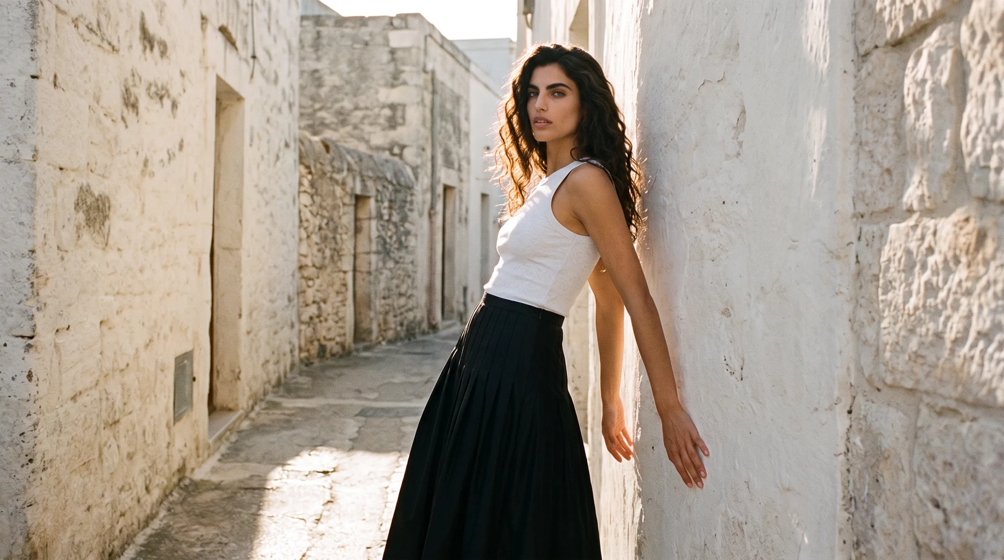 Model in a white tank and black pleated maxi skirt leaning against a sunlit whitewashed wall in a narrow stone lane, late afternoon light