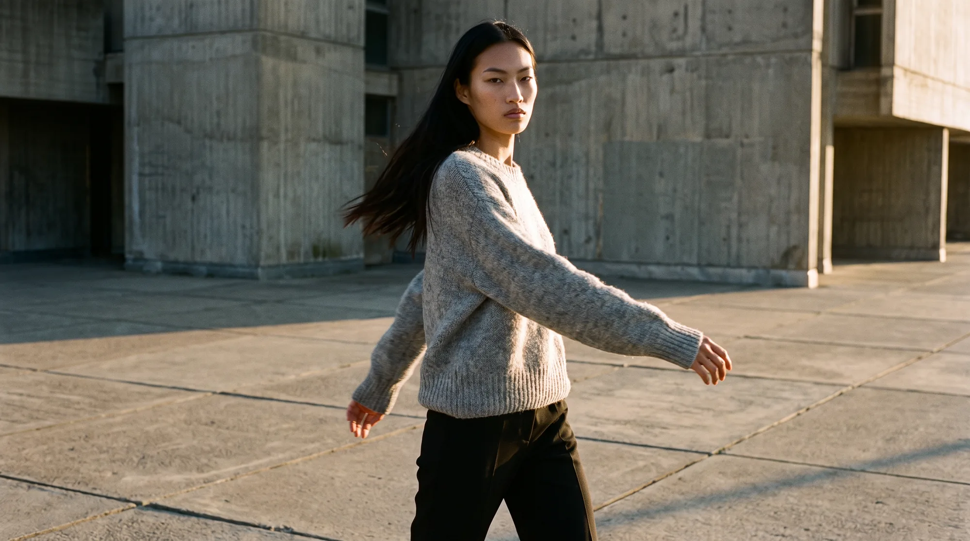 Model in a grey knit sweater and black trousers caught mid-turn on an empty concrete plaza in low raking golden hour light, motion blur in one sleeve and her hair
