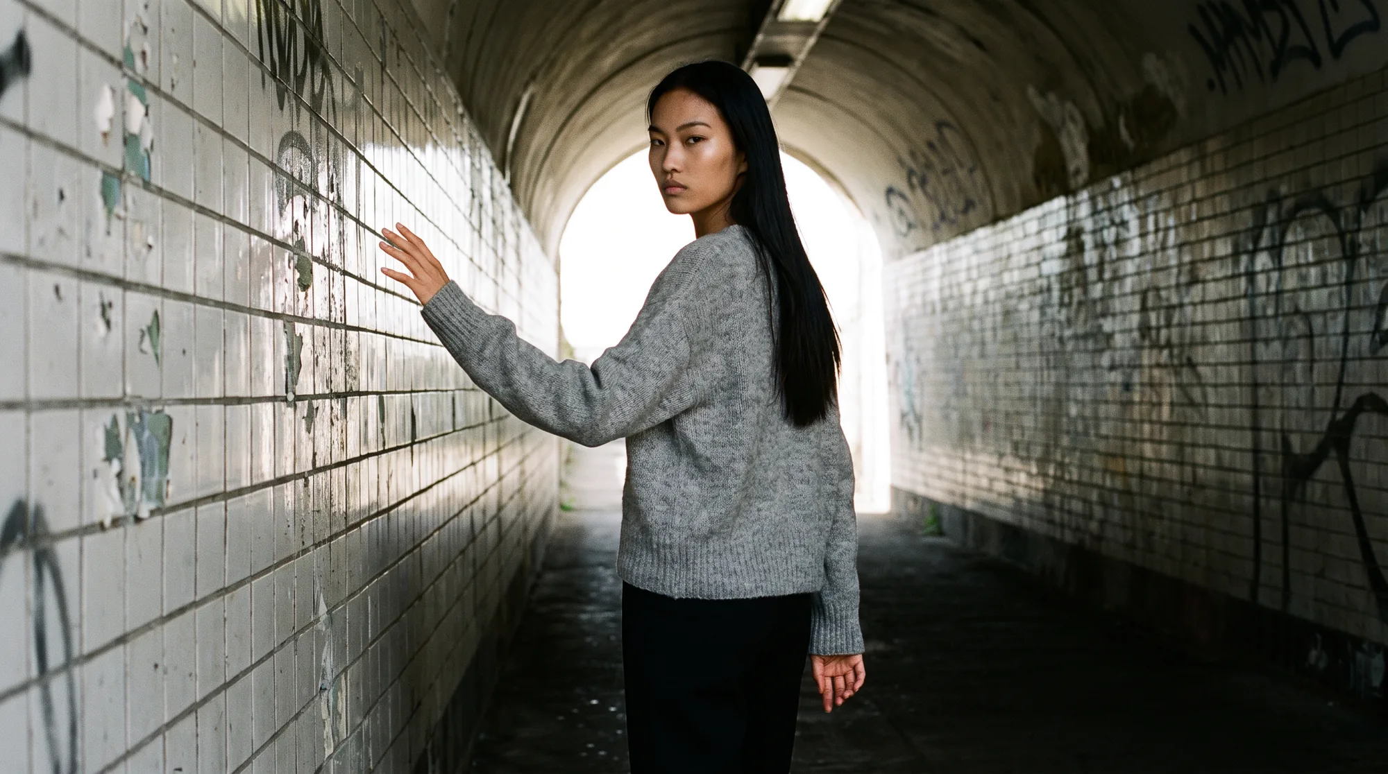 Model in a grey knit sweater and black trousers standing in a tiled underpass with one hand raised to the wall, rim lit against the bright opening at the far end