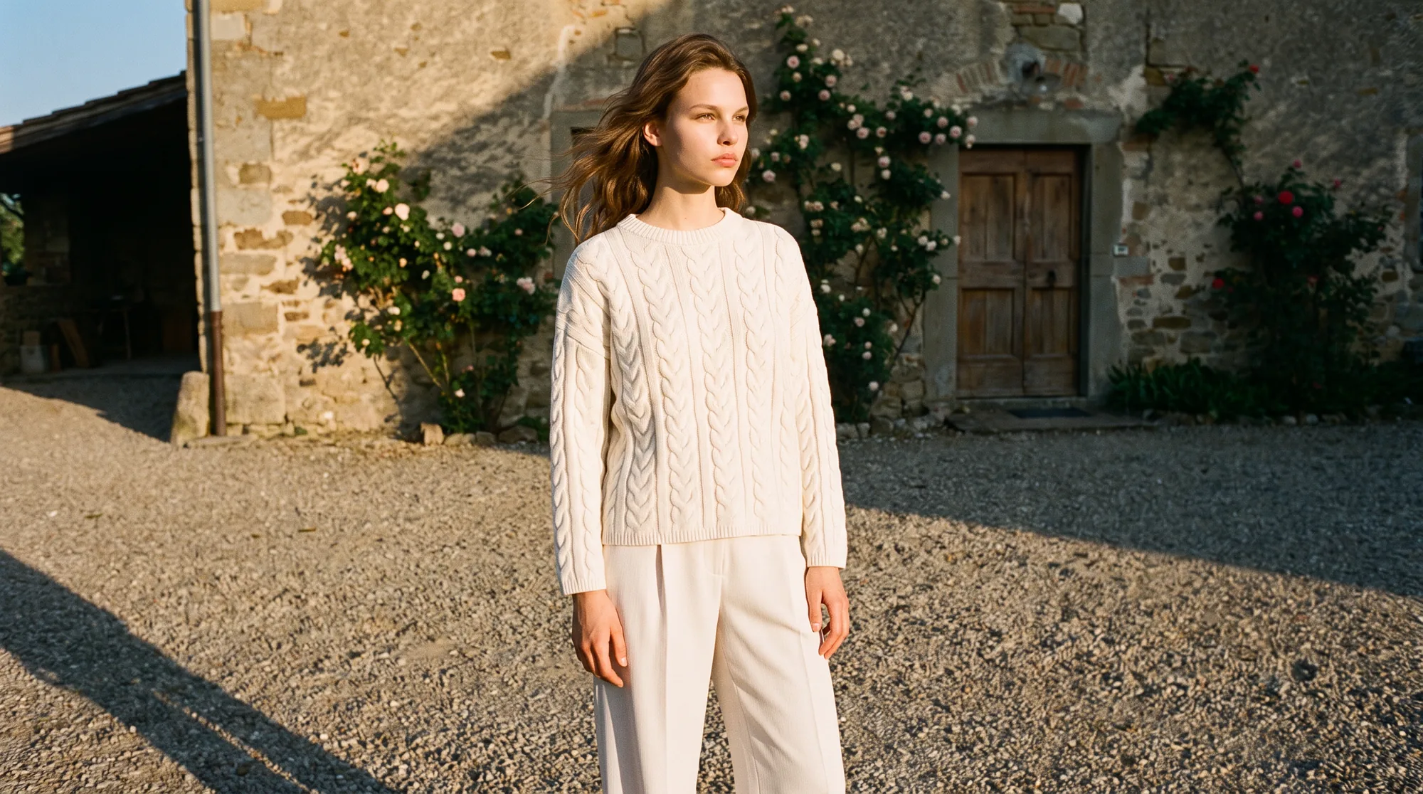 Model in a cream cable knit and off-white wide leg trousers in a gravel courtyard at golden hour
