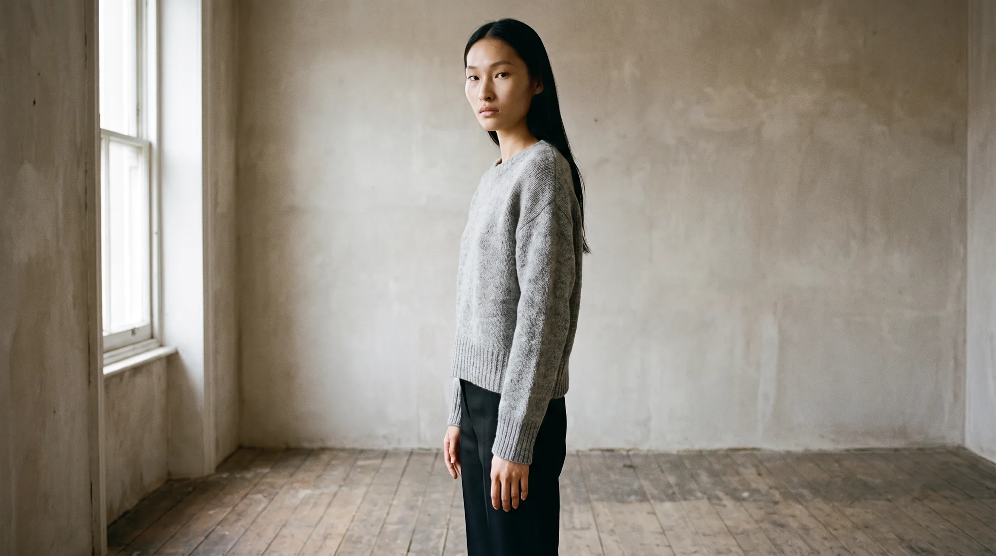 Model in a grey knit sweater and black trousers in a bare plastered room beside a tall window