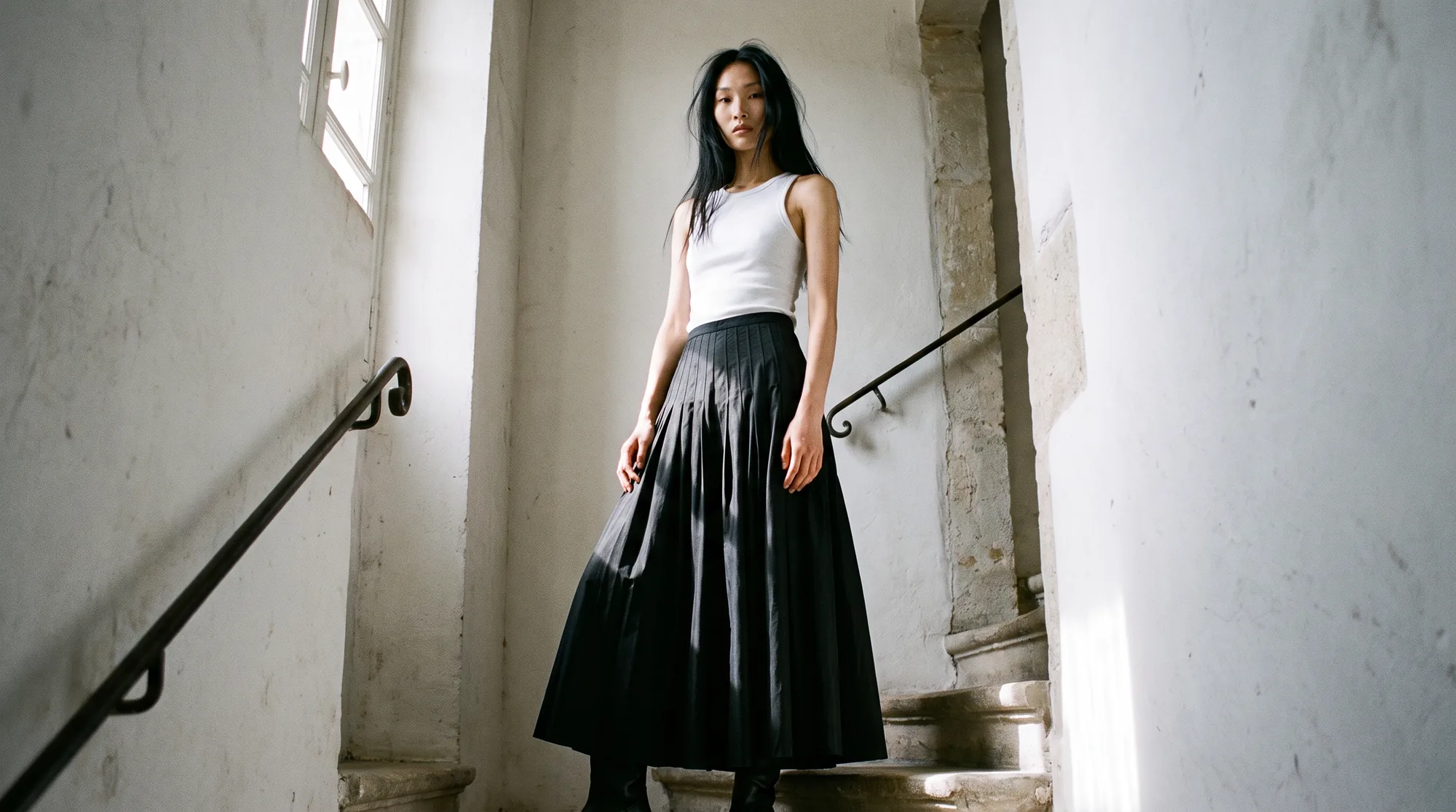 Model in a white top and black pleated skirt on the landing of a whitewashed stone staircase