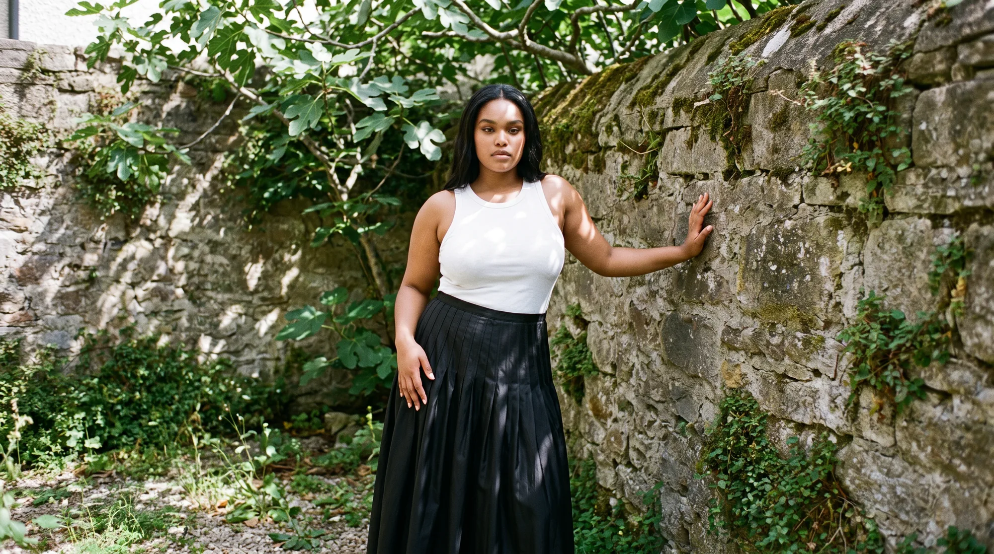 Model in a white top and black pleated skirt in a courtyard garden under a fig tree