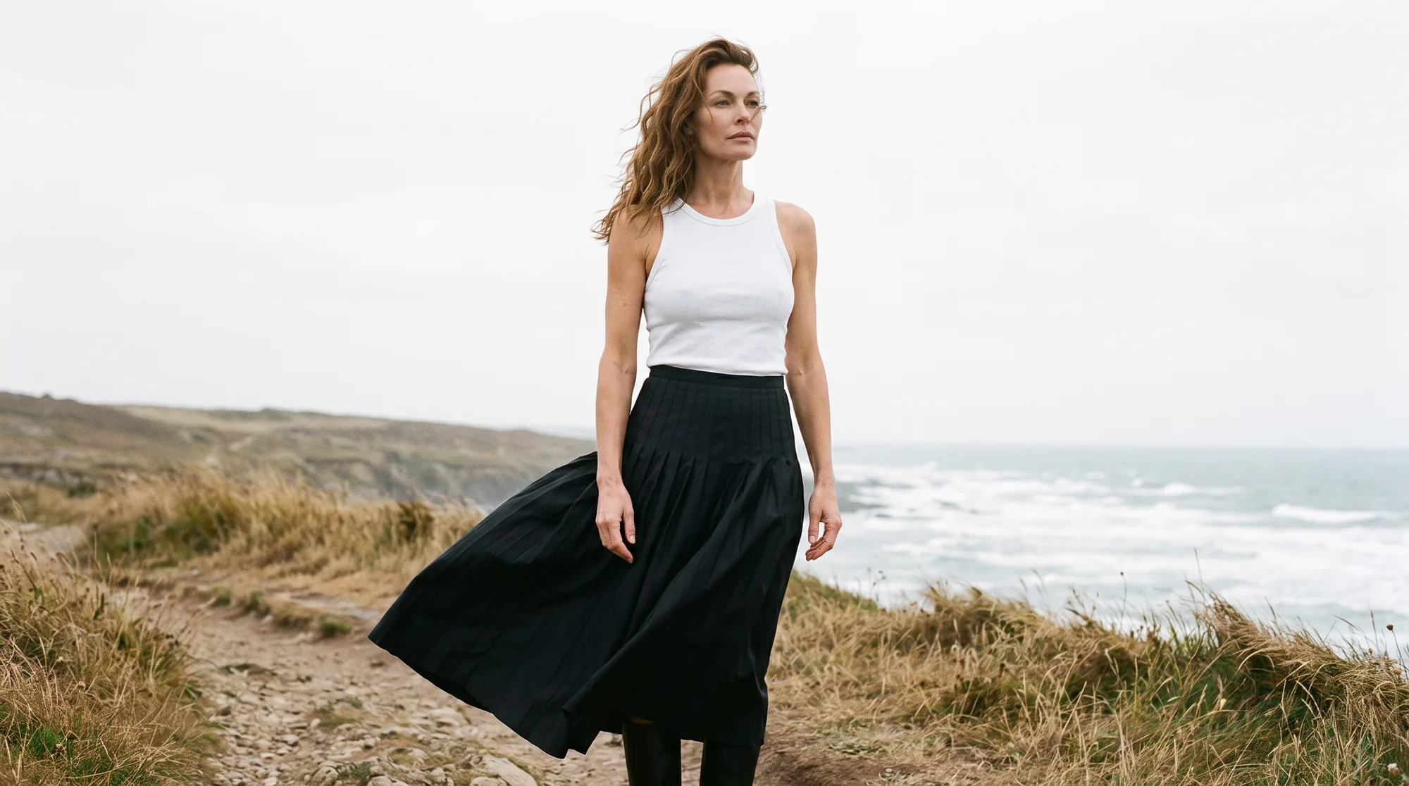 Model in a long pleated skirt on a windswept coastal path under an overcast sky