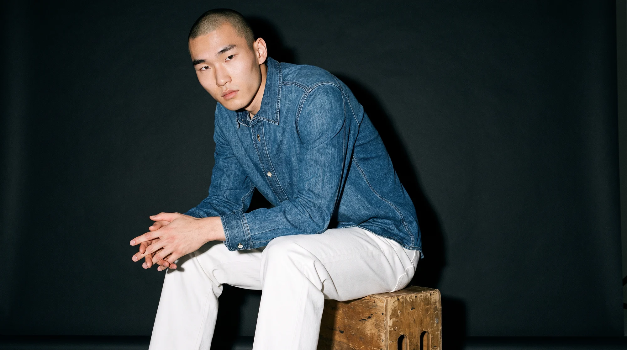 Man in a denim shirt and white jeans seated on a wooden box against a charcoal backdrop, AI on-model image