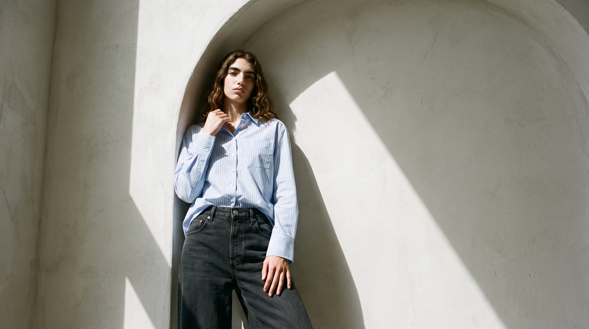 Woman in a pale blue striped shirt and black wide-leg jeans leaning in a sunlit plaster alcove, the same AI model in a different setting