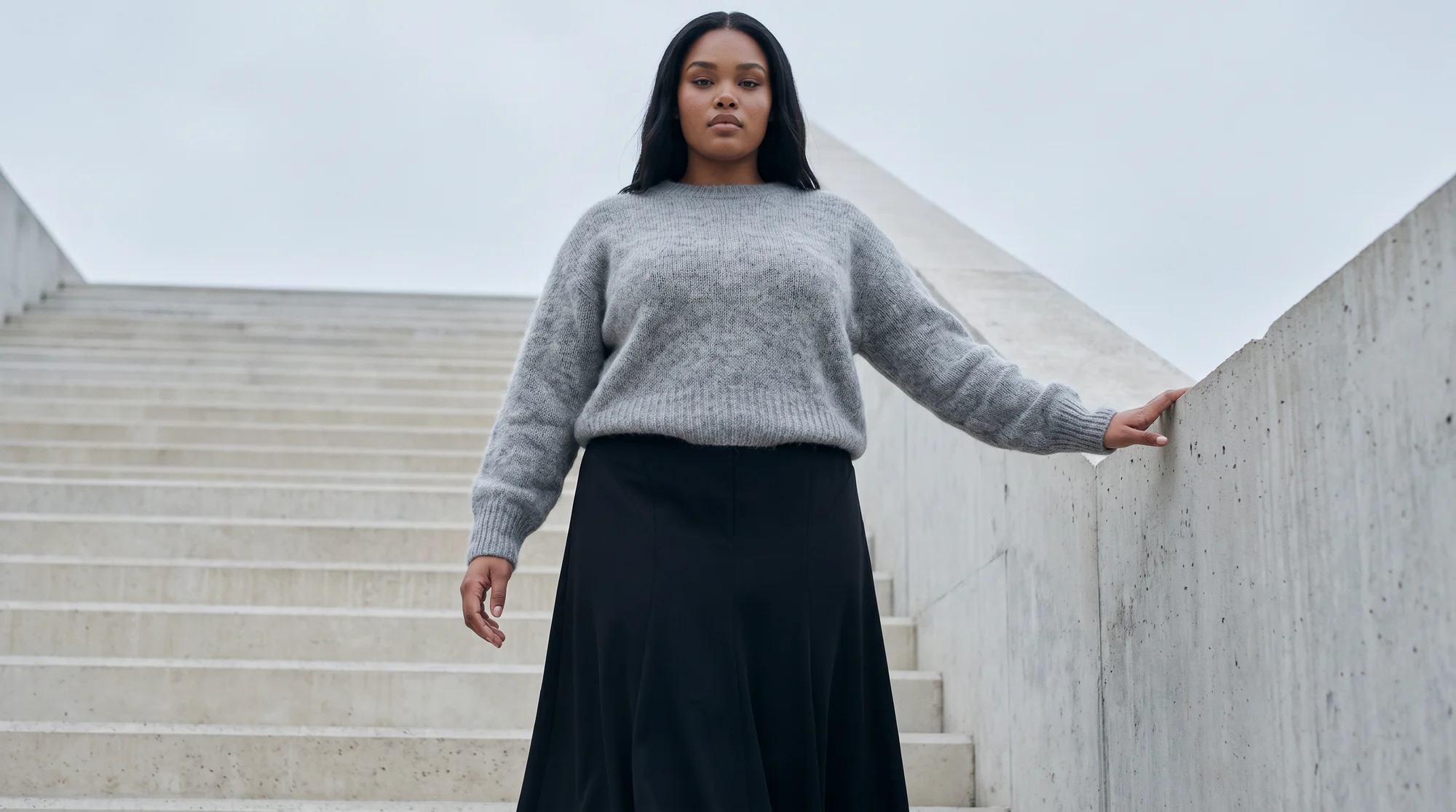 Woman in a grey mohair sweater and black skirt on wide concrete steps with one hand on the wall, AI lookbook image