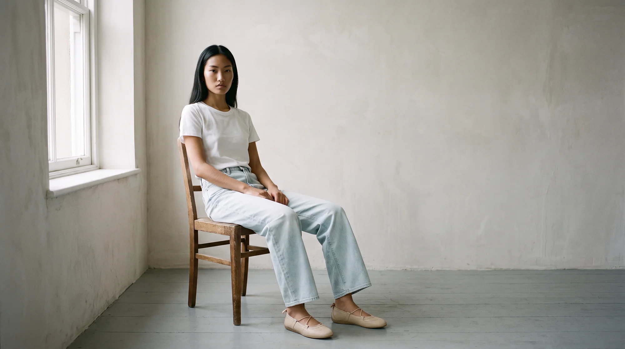 Woman in a white t-shirt and pale jeans seated on a wooden chair beside a window in a plaster-walled room, AI fashion model