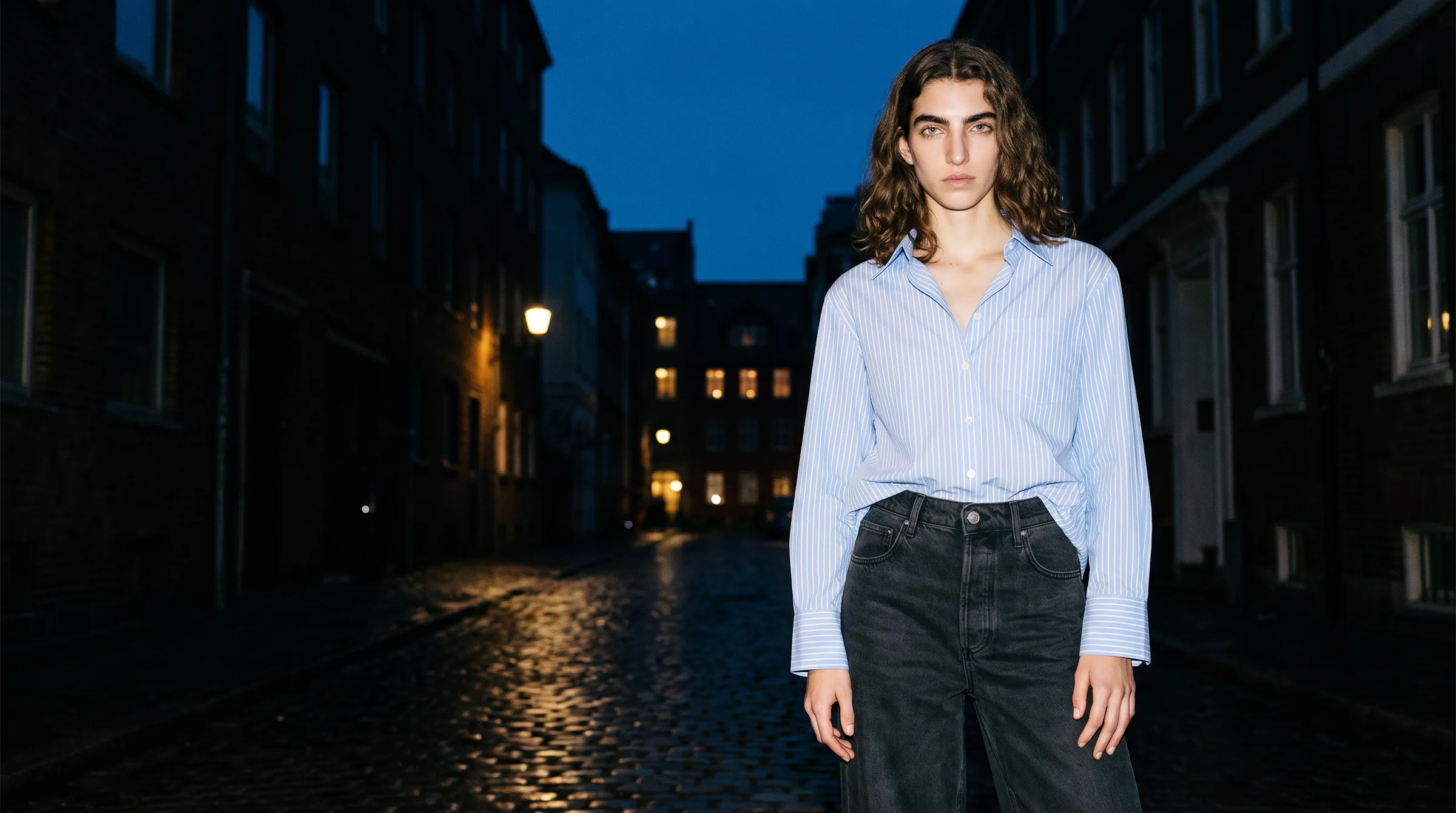 AI fashion model in a pale blue striped shirt and black jeans on an empty street at dusk lit by hard on-camera flash