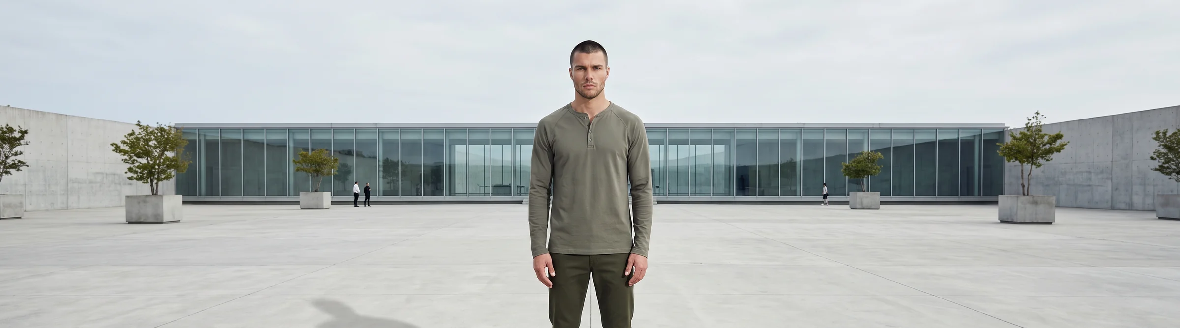 Male AI fashion model in an olive henley standing in front of a long low glass building, cropped from the knees up