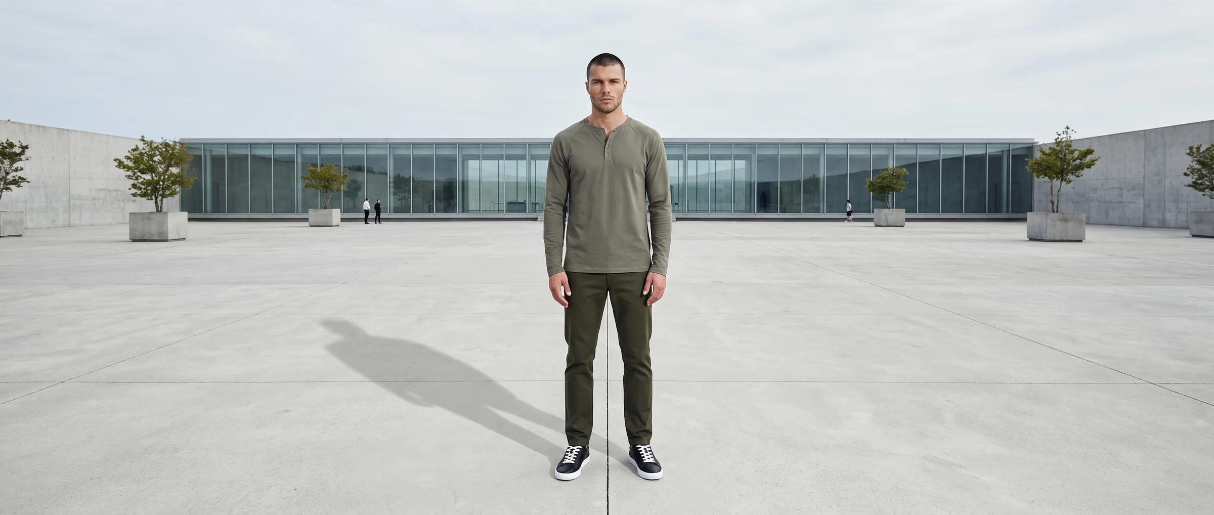 Male AI fashion model in an olive henley and trousers standing alone in the centre of a vast concrete forecourt in front of a long low glass building