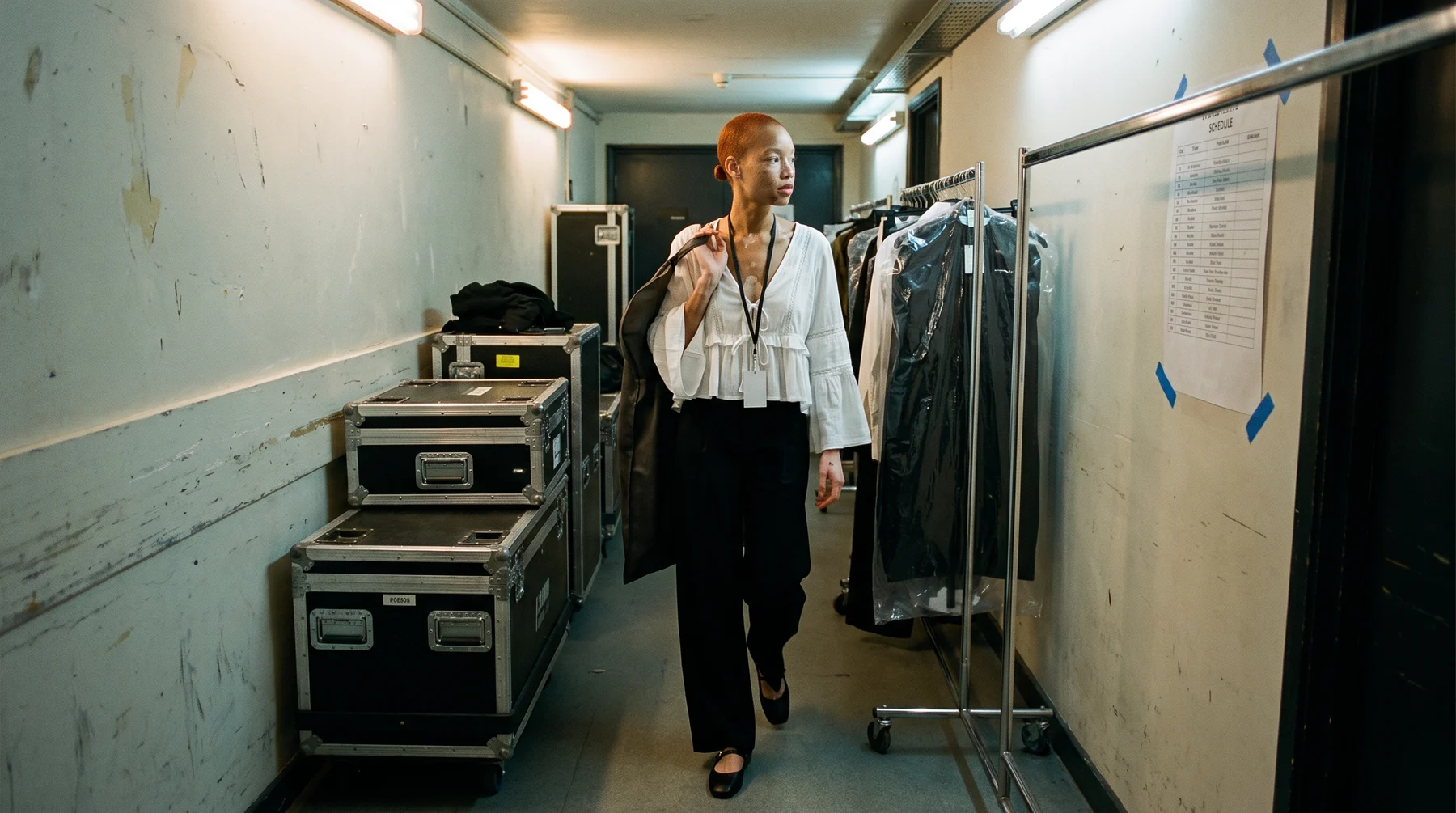 Woman in a white blouse carrying a garment bag along a backstage corridor past flight cases and a rail of garment bags
