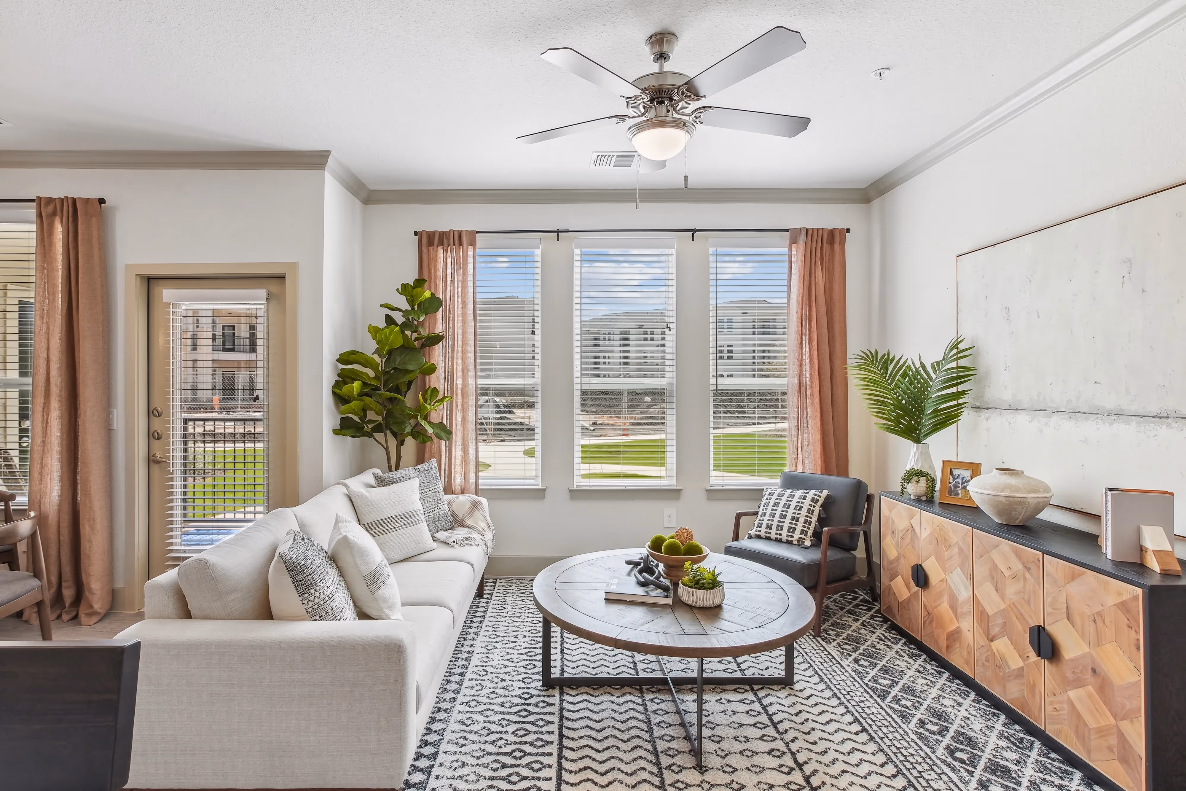 Spacious living room with a white sofa, ceiling fan, and large windows with blinds and curtains, opening to a balcony at Paloma Luxury Apartments in San Antonio, TX.