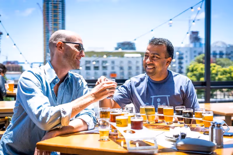Two men smiling and toasting beer flights at an outdoor brewery, representing the local dining and social scene near Paloma Luxury Apartments in San Antonio, TX.