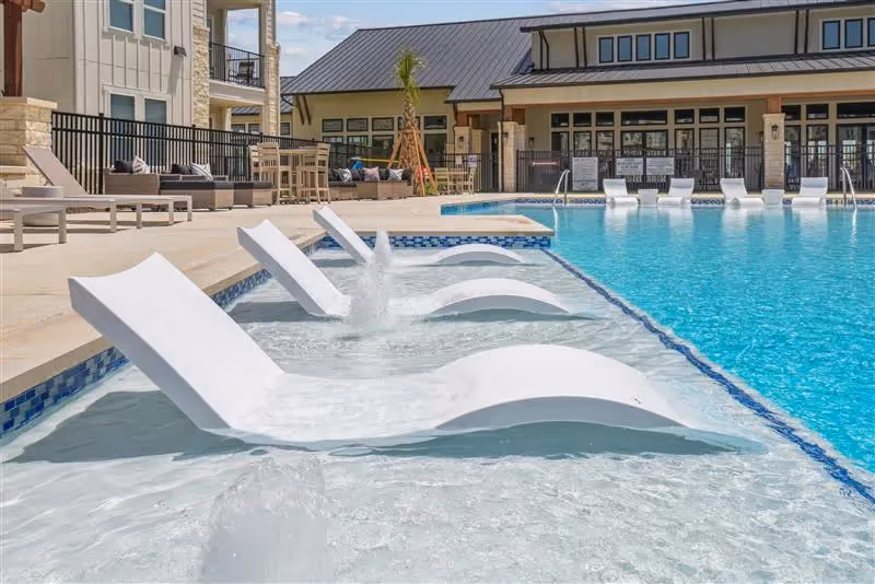 Resort-style swimming pool with in-pool loungers and a modern clubhouse in the background, a key amenity at Paloma apartments in San Antonio, TX.