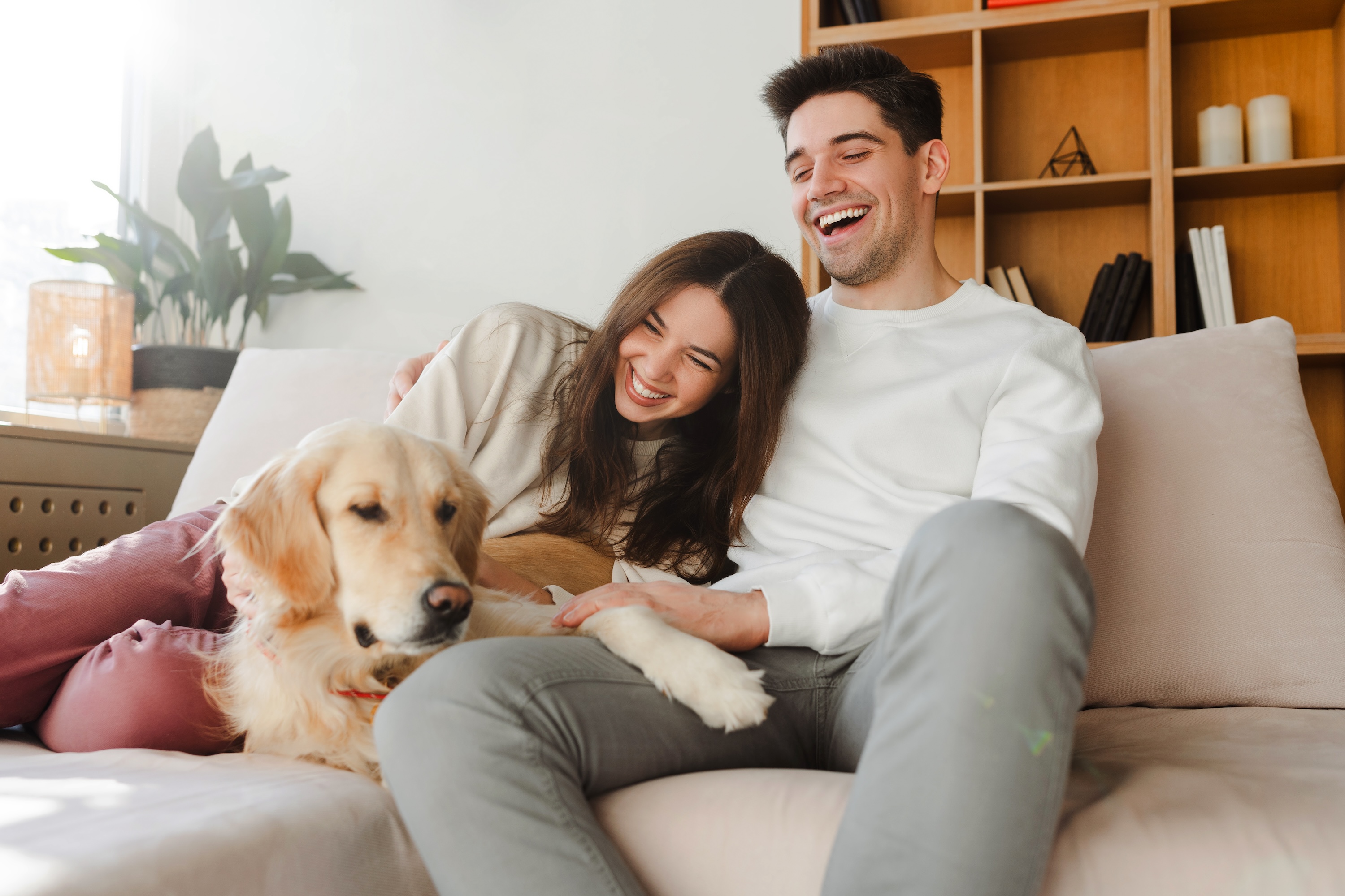 A happy couple laughing on a sofa with their golden retriever dog, representing the pet-friendly and joyful home life at Paloma in San Antonio, TX.