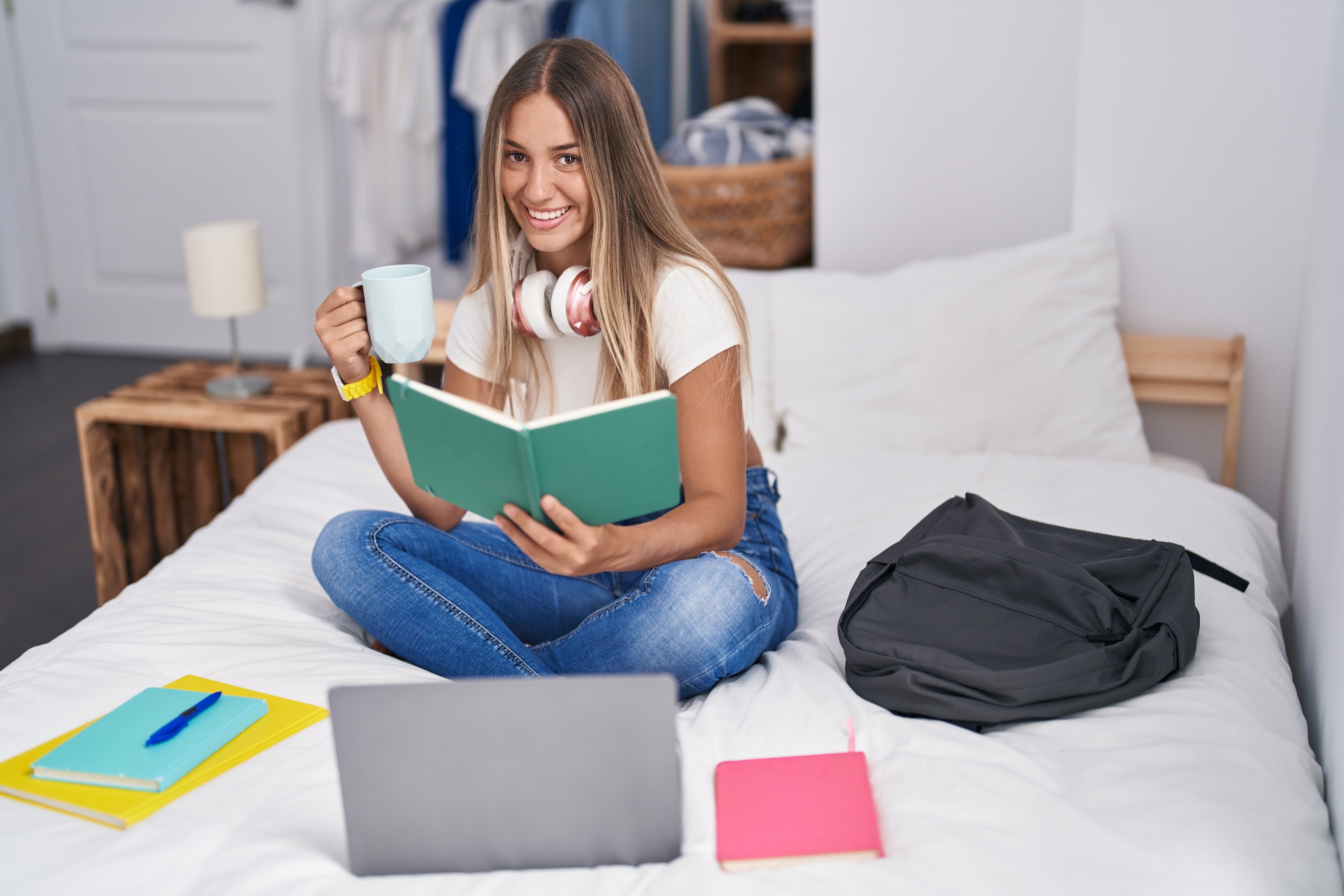 A resident sitting cross-legged on a luxurious, clean white bed with a coffee mug and a book, a laptop nearby, highlighting the comfort and spaciousness of the apartment bedrooms.