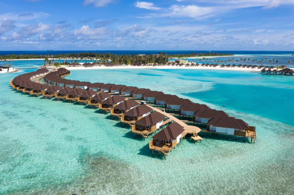 Overwater villas with brown roofs extending in a curved line over the lagoon at a Maldives resort, with white sandy beaches and palm trees in the background.
