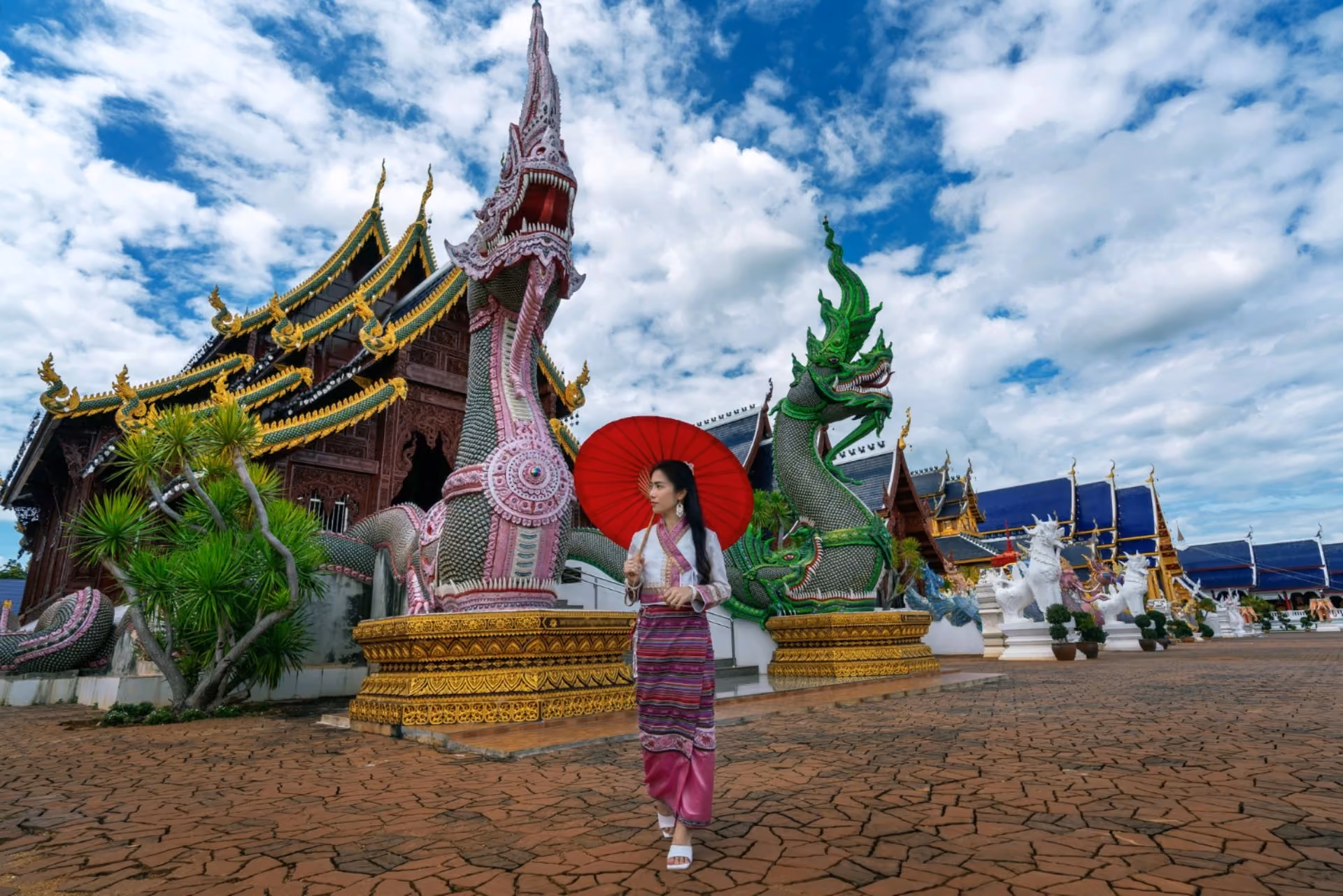 Woman in traditional Thai dress holding red umbrella at ornate Naga temple – Bali vs Thailand cultural experience