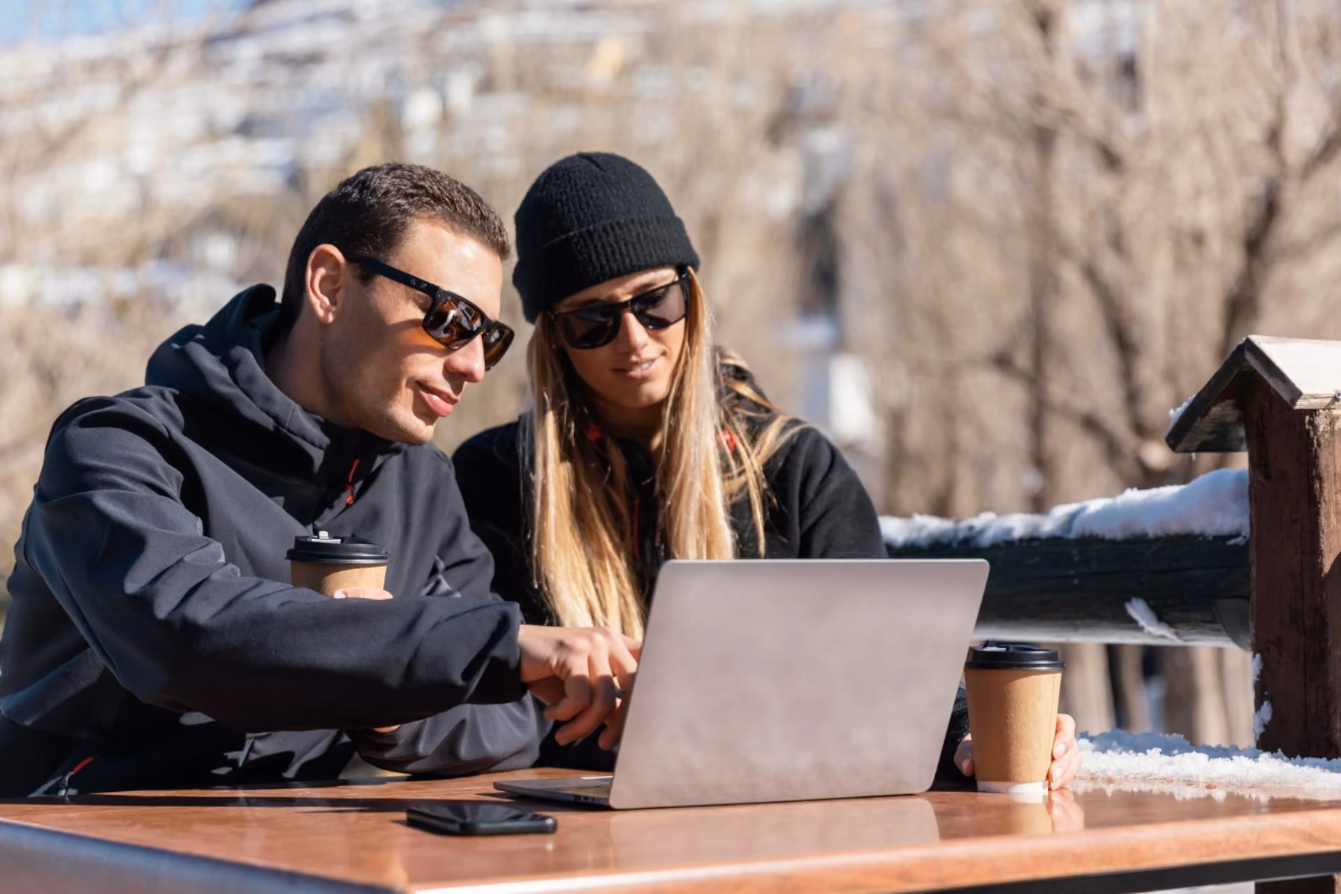 Couple working outdoors with coffee and laptop during winter travel, comparing New Zealand vs Switzerland for peaceful romantic escapes