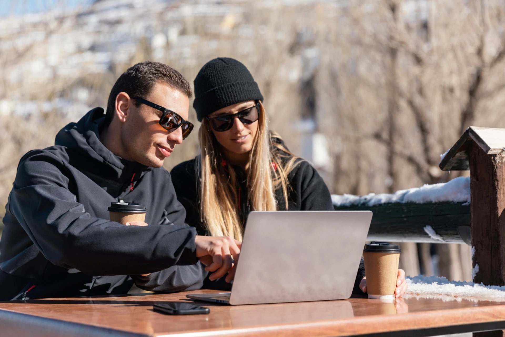 Couple working outdoors with coffee and laptop during winter travel, comparing New Zealand vs Switzerland for peaceful romantic escapes