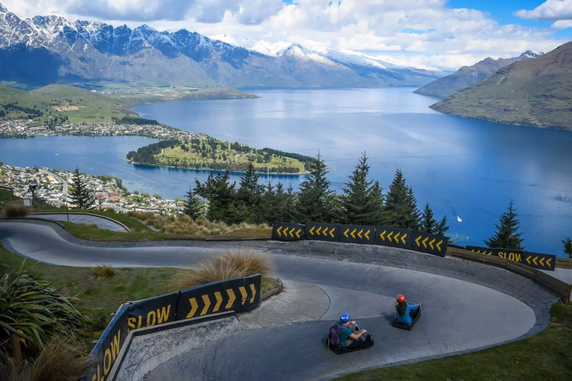 Luge riders racing down track overlooking New Zealand lake