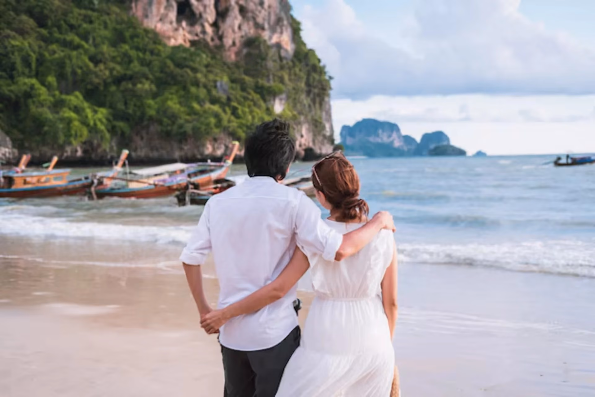Couple holding hands on Krabi beach with boats and limestone cliffs