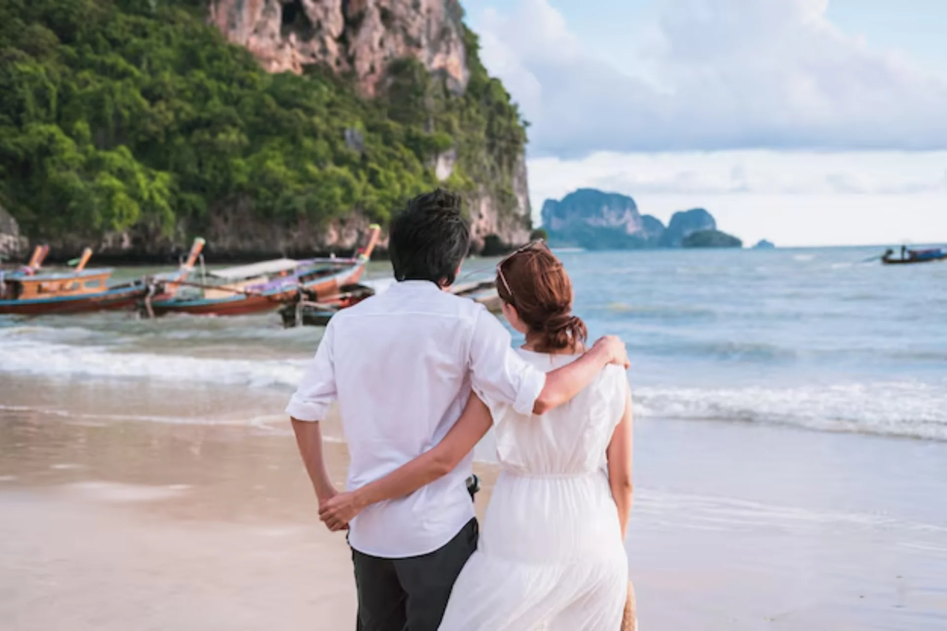 Couple holding hands on Krabi beach with boats and limestone cliffs