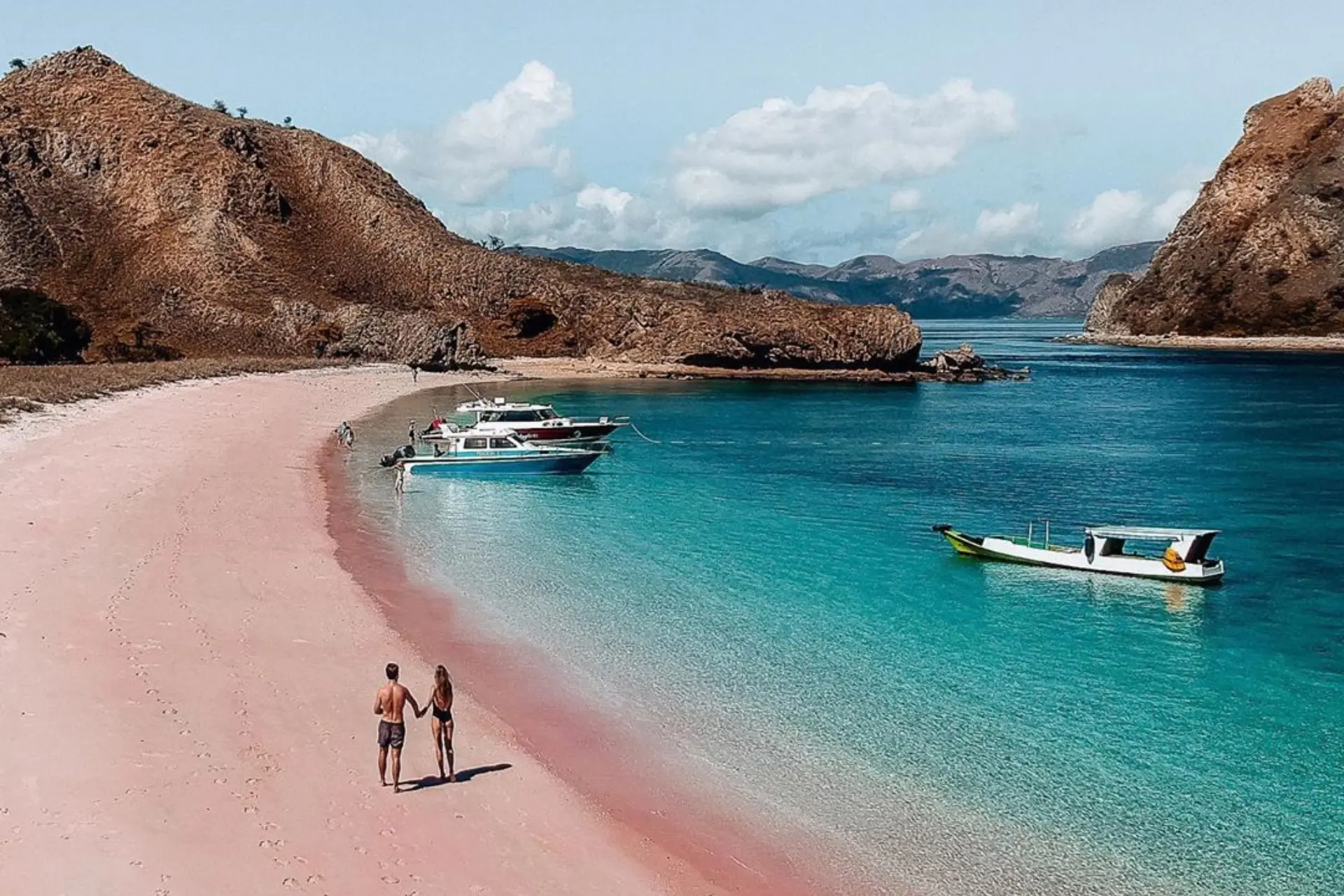 Couple wading in pink sand beach Lombok with boats